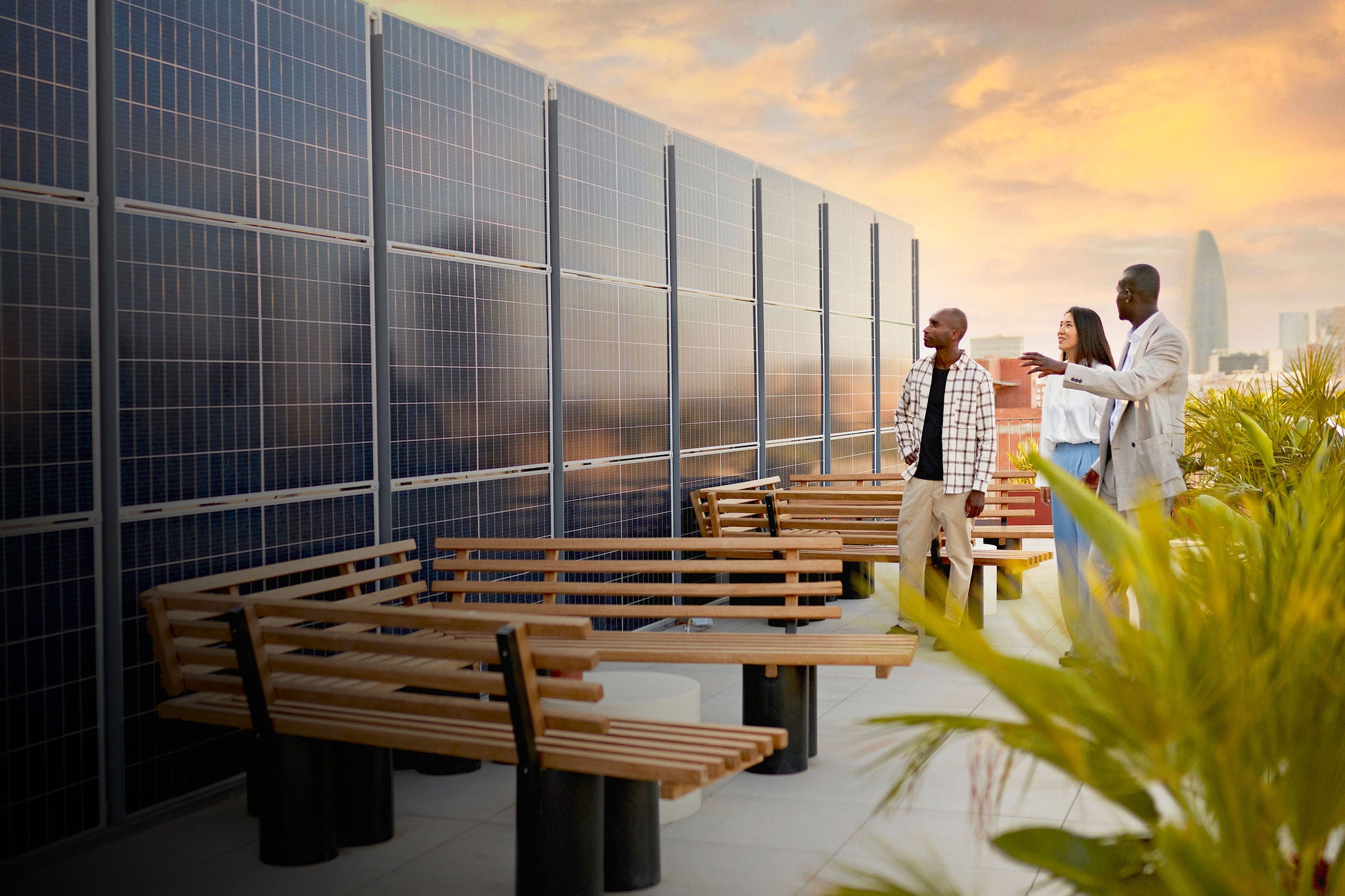Real estate agent and couple standing on rooftop of environmentally aware office building with dramatic sky and Barcelona cityscape in background at sunset.