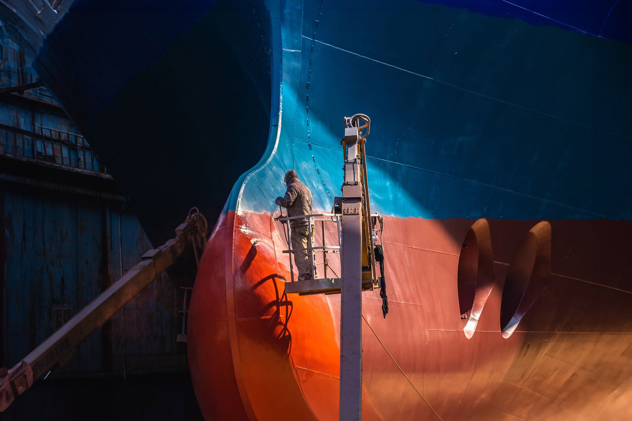 Ein Arbeiter steht auf einer Hebebühne und lackiert den großen Rumpf eines Schiffes in der Werft bei Lichtim Hafen.