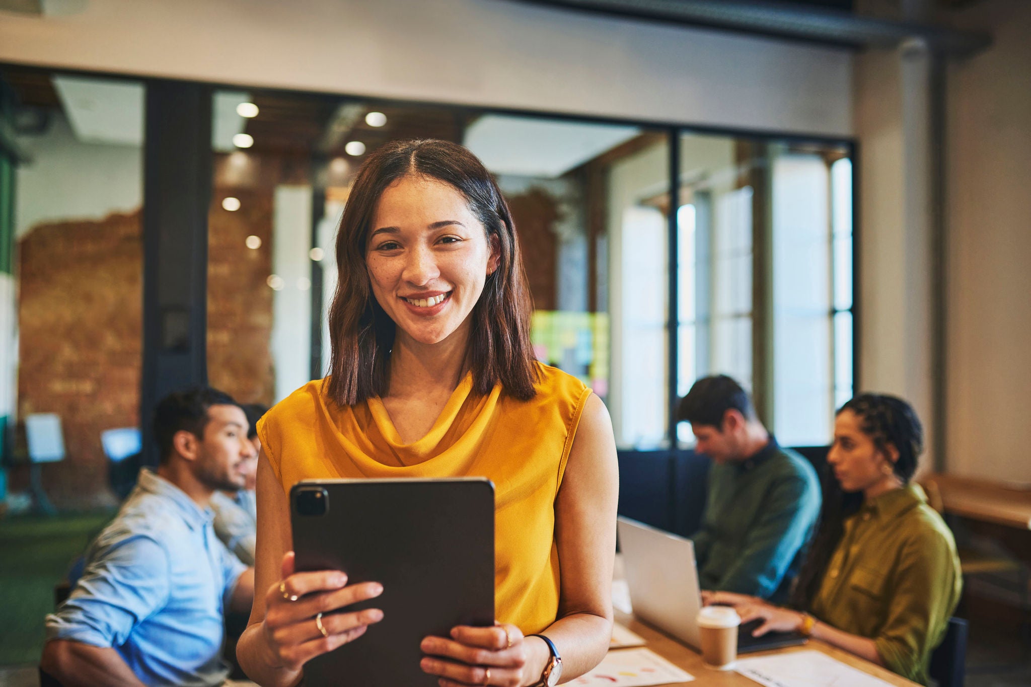 Businesswoman smiling cheerfully at camera, holding digital tablet in meeting room with colleagues at discussion in background