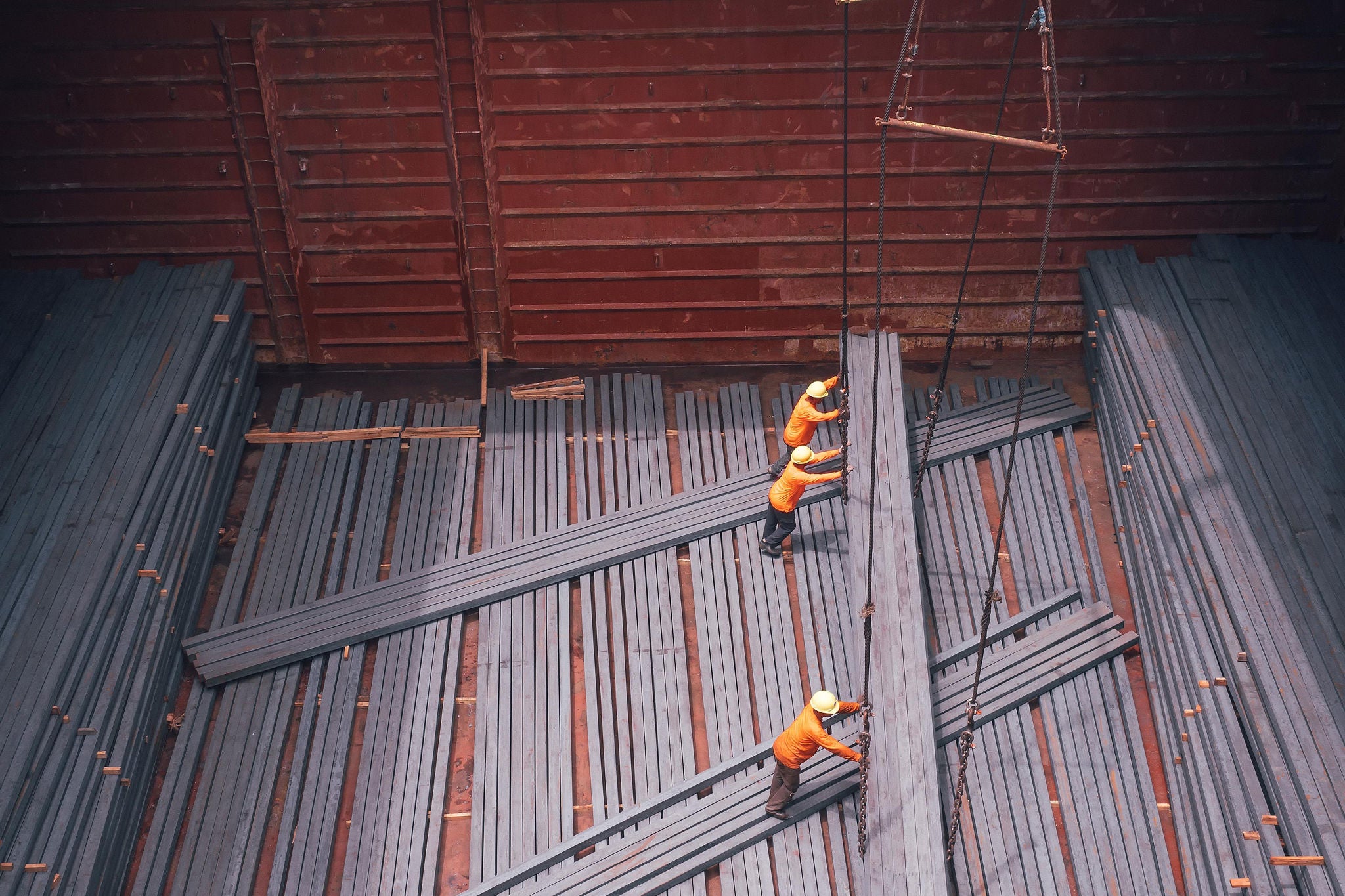 Workers moving long metal beams inside a cargo hold viewed from above