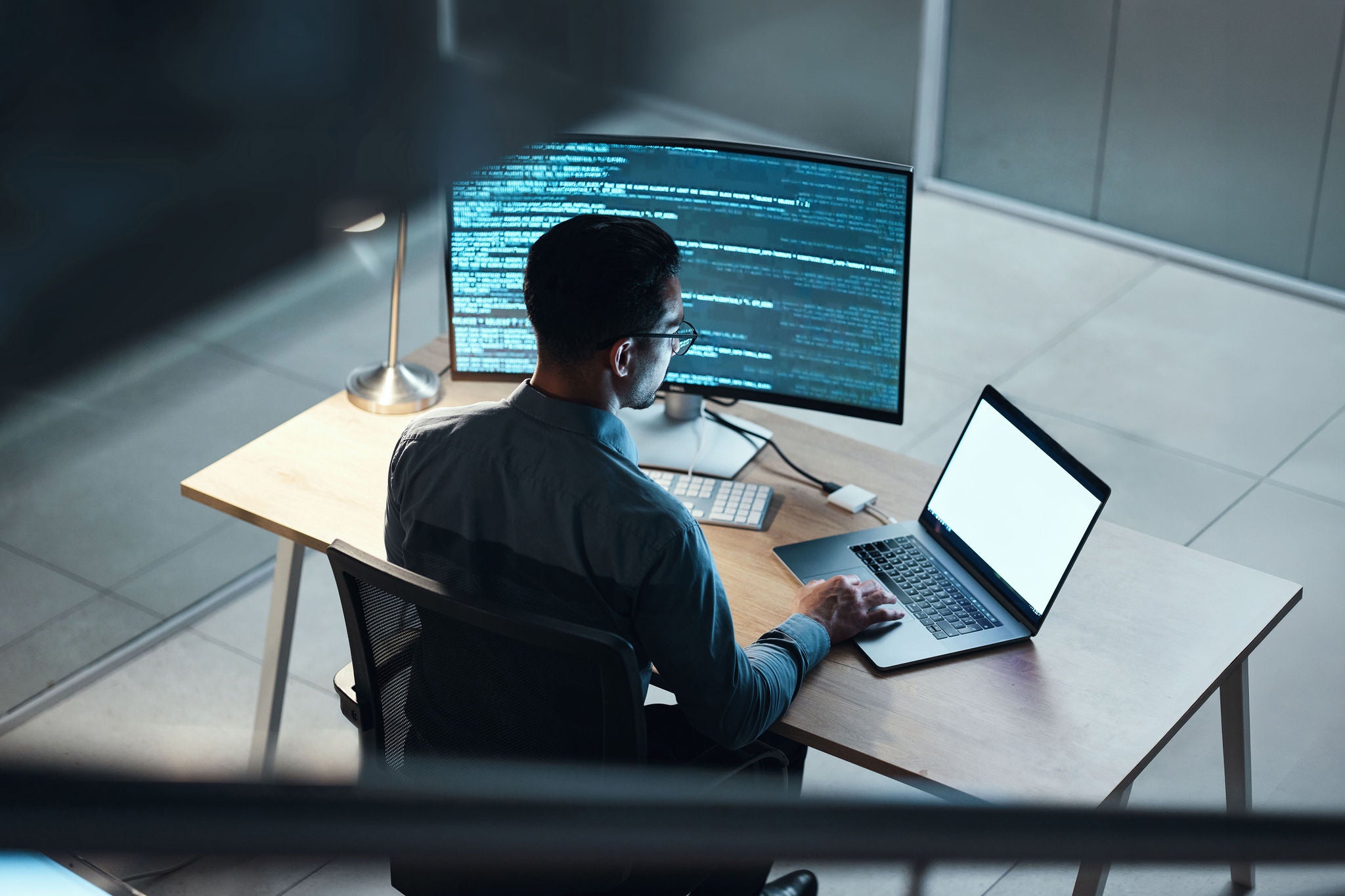 Eine Person sitzt an modernem Arbeitsplatz mit großem Monitor, Laptop und sichtbarem ProgrammcodeIT