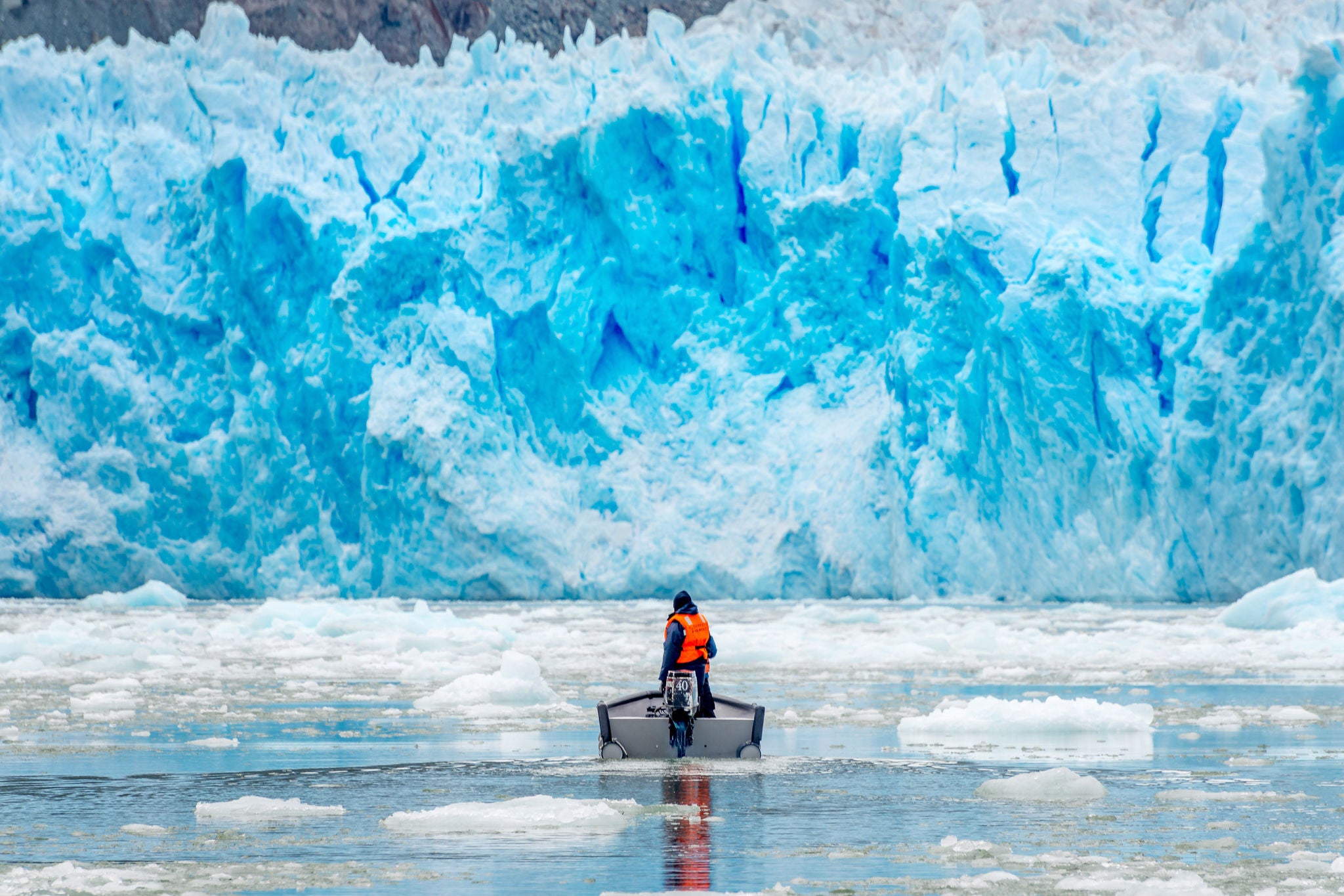 Person in a small boat moving through icy water with a towering blue glacier rising in the background