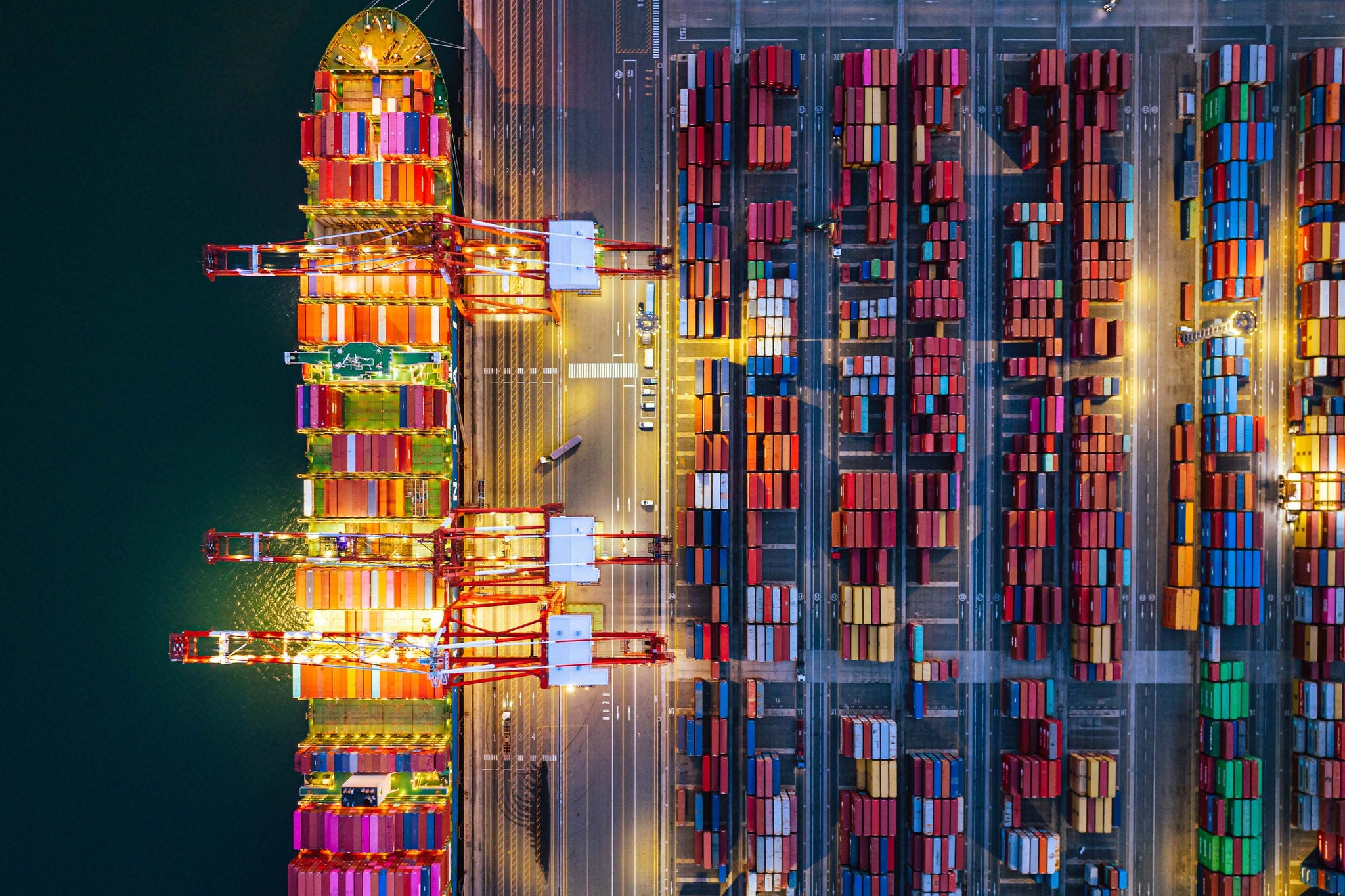 Bunte Containerreihen und beleuchtete Kräne zeigen einen geschäftigen Hafen bei Nacht.