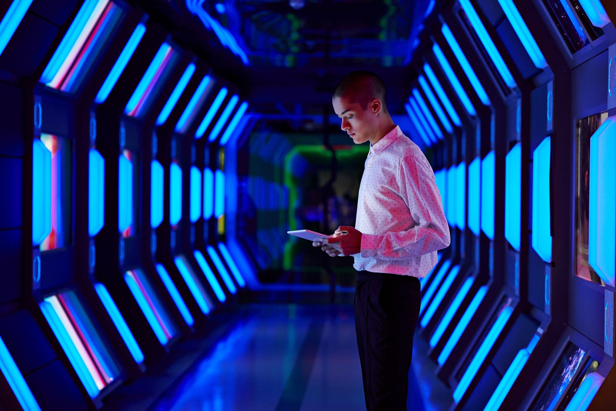 A person holding a tablet stands in a futuristic corridor illuminated by bright blue and purple lights.