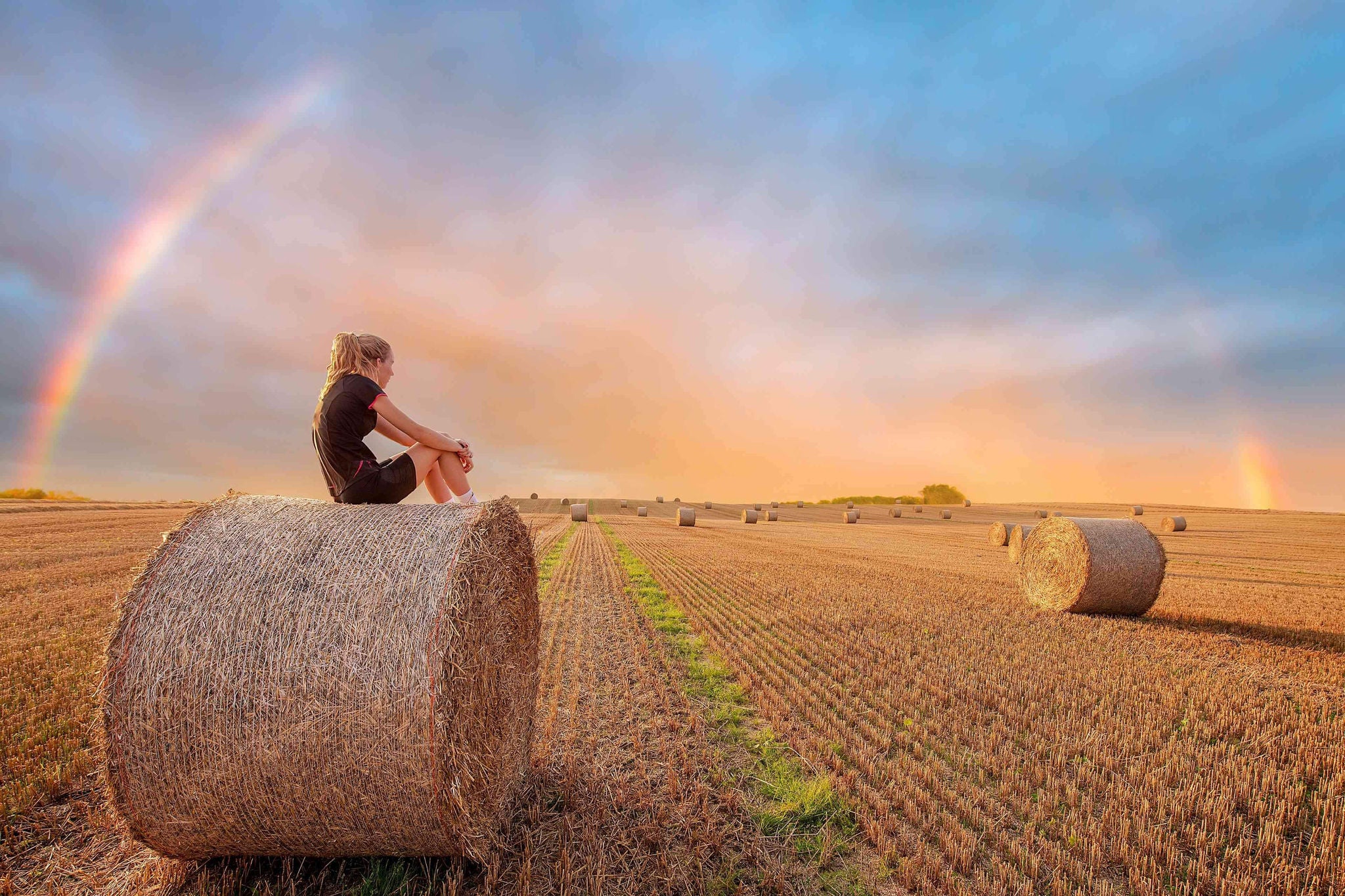 Woman looking sat on hay Bales as a rainbow appears after a rain shower
