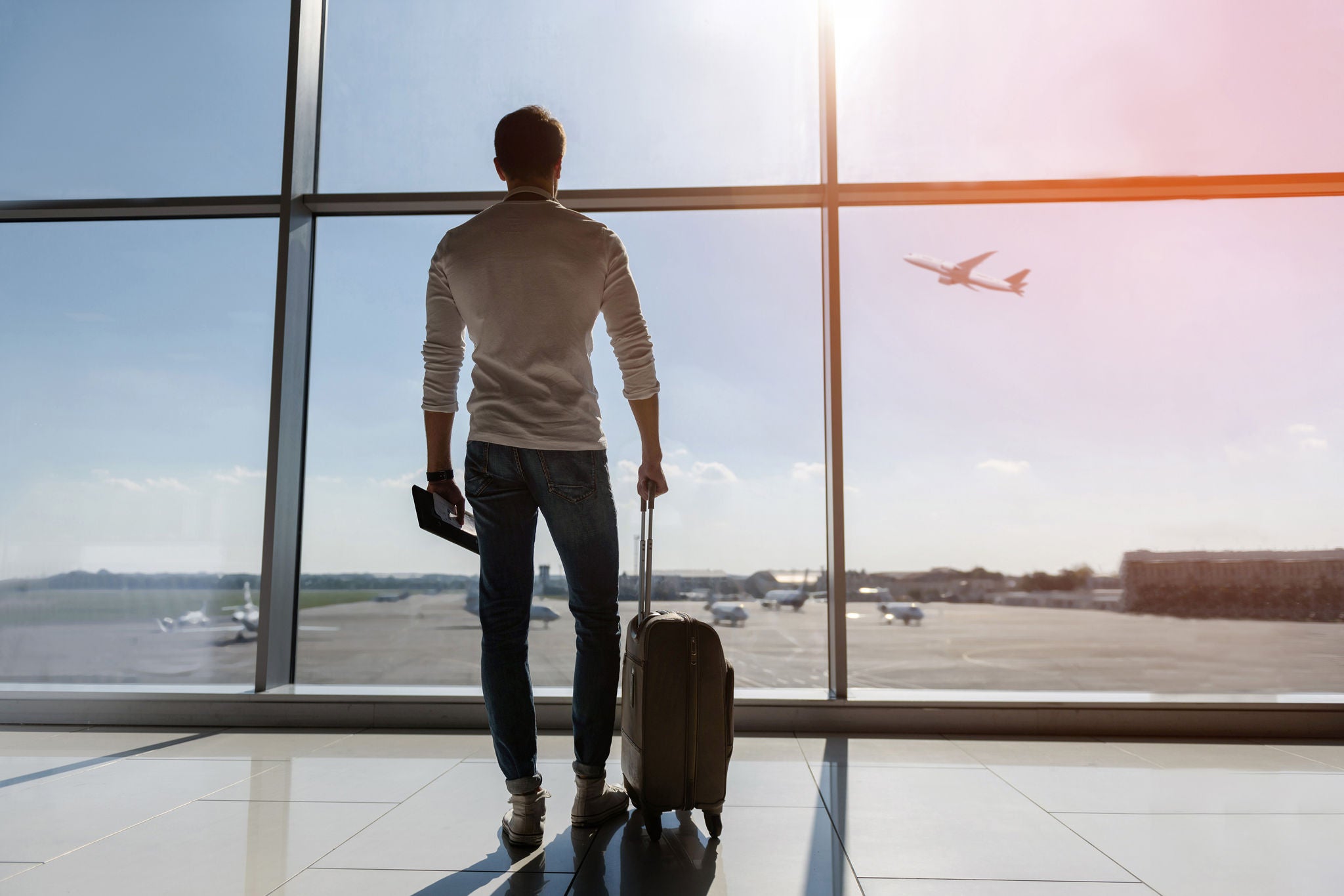 Man stands by an airport window with a suitcase, watching a plane on the runway.