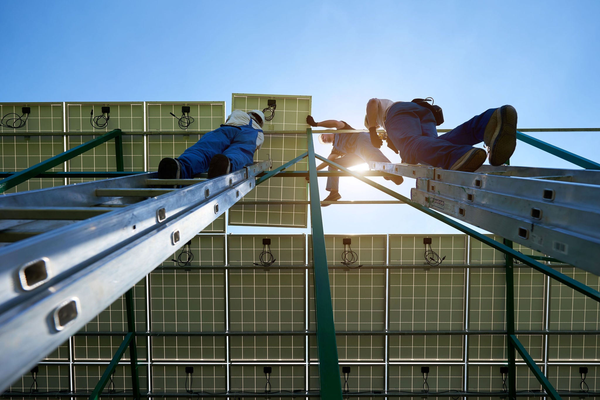 Workers on ladders installing or inspecting solar panels against a clear blue sky.