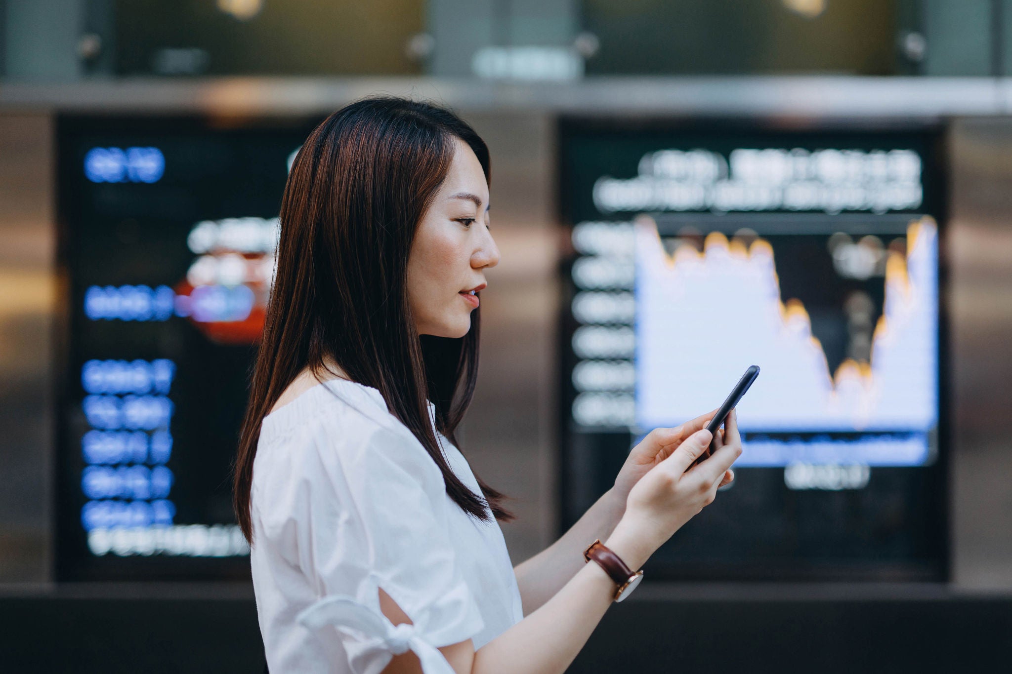 Young Asian businesswoman using smartphone against stock exchange market display screen board in downtown financial district