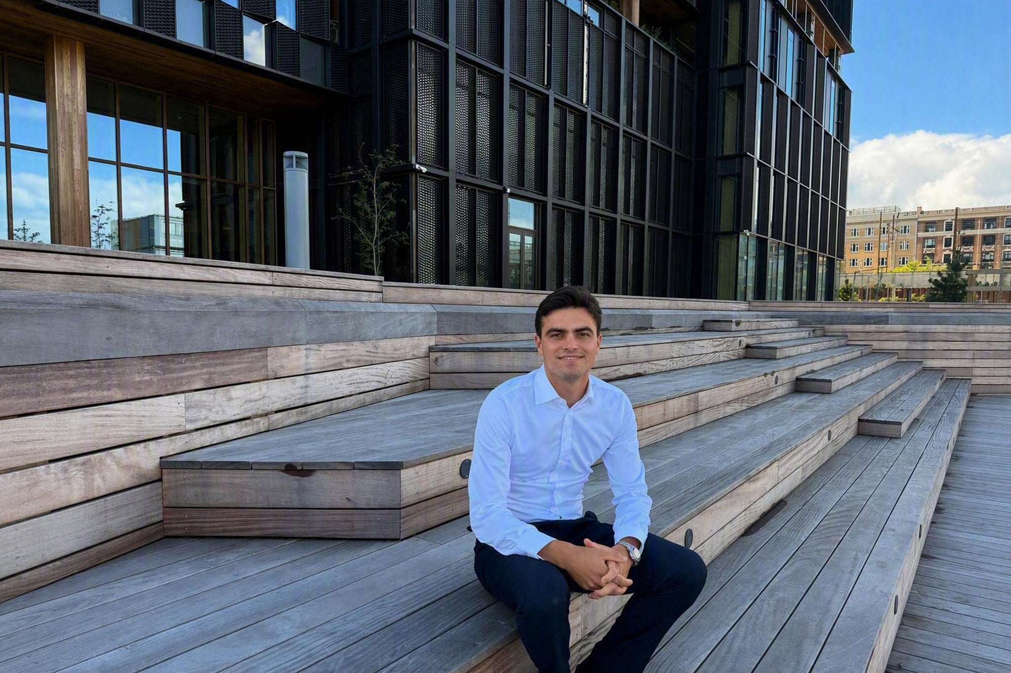 Professional seated on wooden steps outside a modern office building with glass facades and urban surroundings.