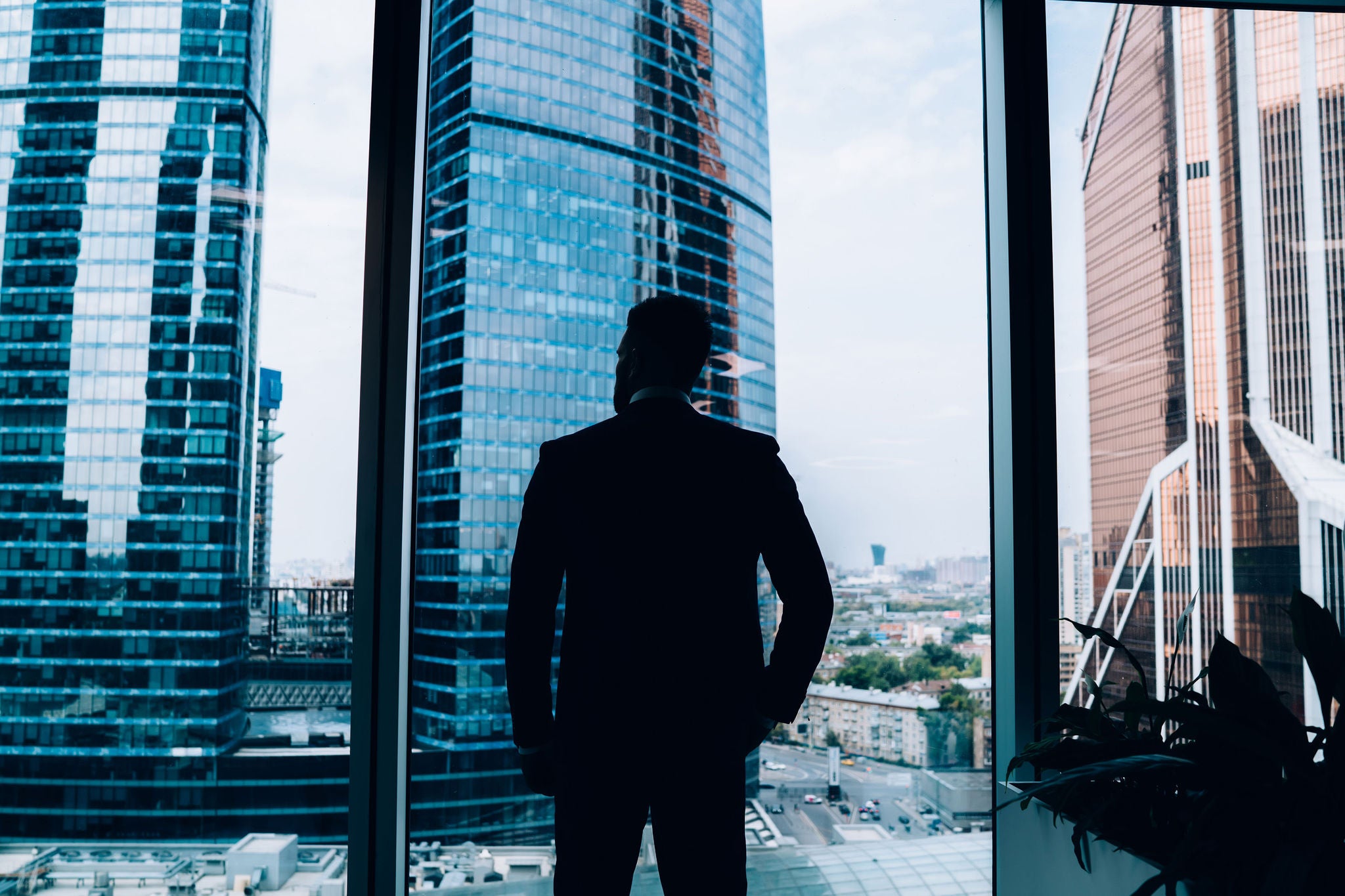Back view of silhouette of successful thoughtful businessman making decision while looking at downtown from panoramic window in modern office