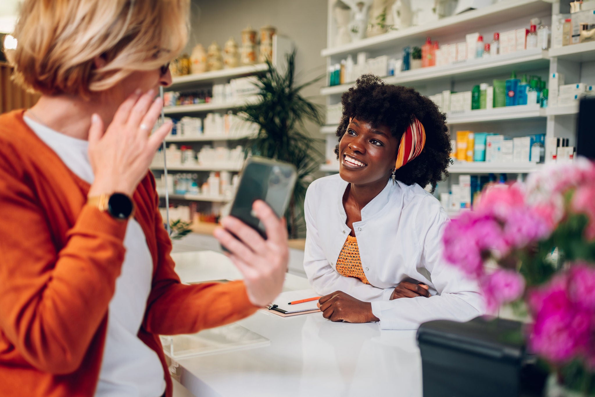 Smiling african american woman healthcare worker working in a modern pharmacy. Confident pharmacist consulting a senior customer about a medicine and looking into her smartphone. Copy space. Diversity