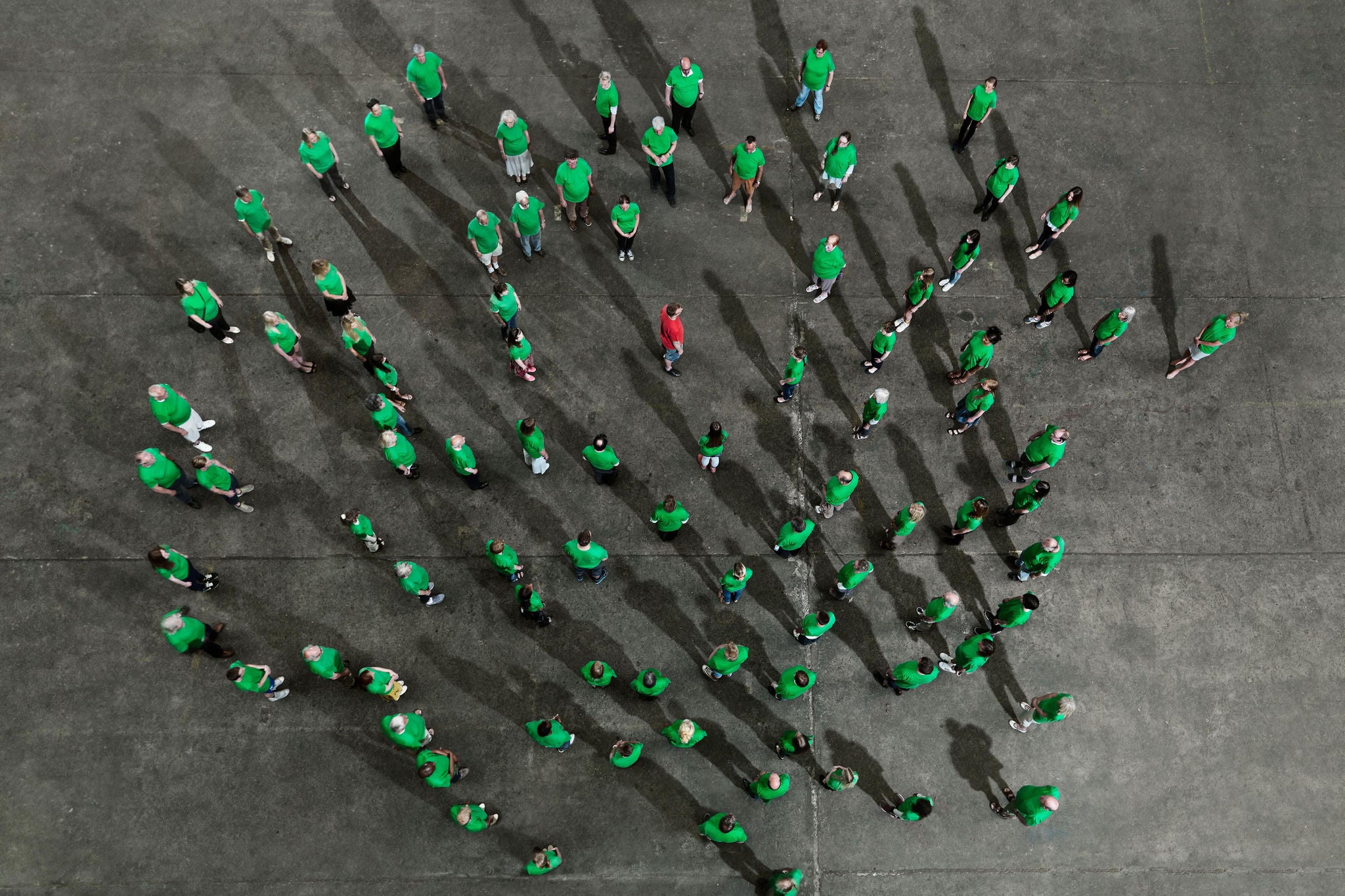 A crowd of people wearing coloured looking towards a person standing in the middle of the group, wearing a red shirt. Photographed from above.