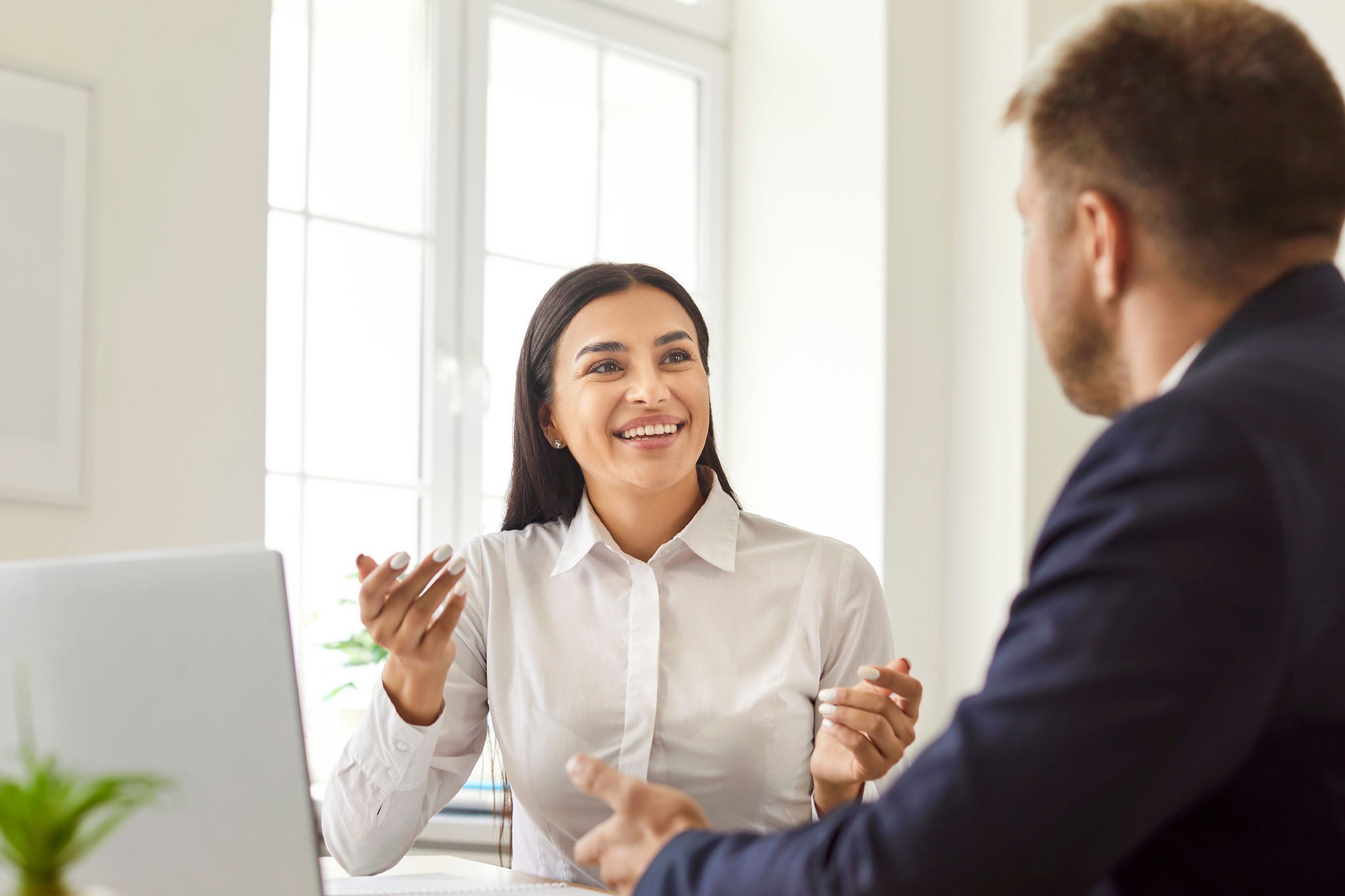 Professional financial advisor talking to a client. Happy smiling friendly young brunette woman in a white shirt sitting at an office desk, communicating with a male customer, offering a business deal