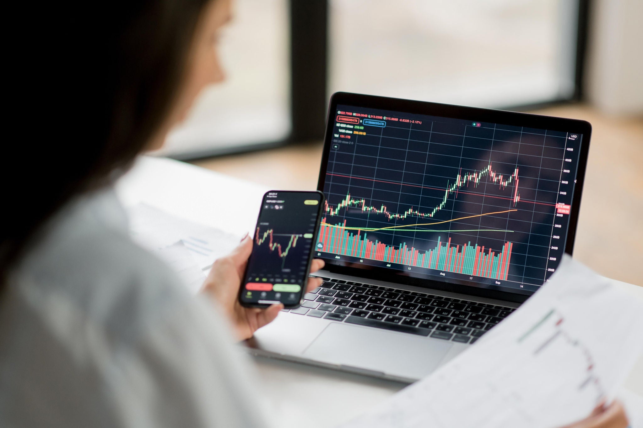 Person viewing stock price charts on a laptop and smartphone at a desk.