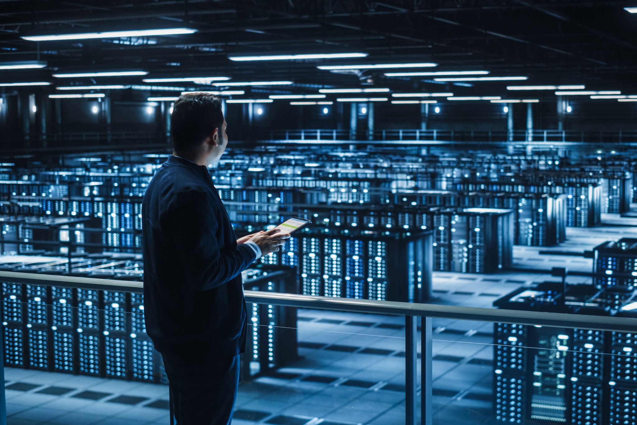 Handsome Smiling IT Specialist Using Tablet Computer in Data Center. Succesful Businessman and e-Business Entrepreneur Overlooking Server Farm Cloud Computing Facility.