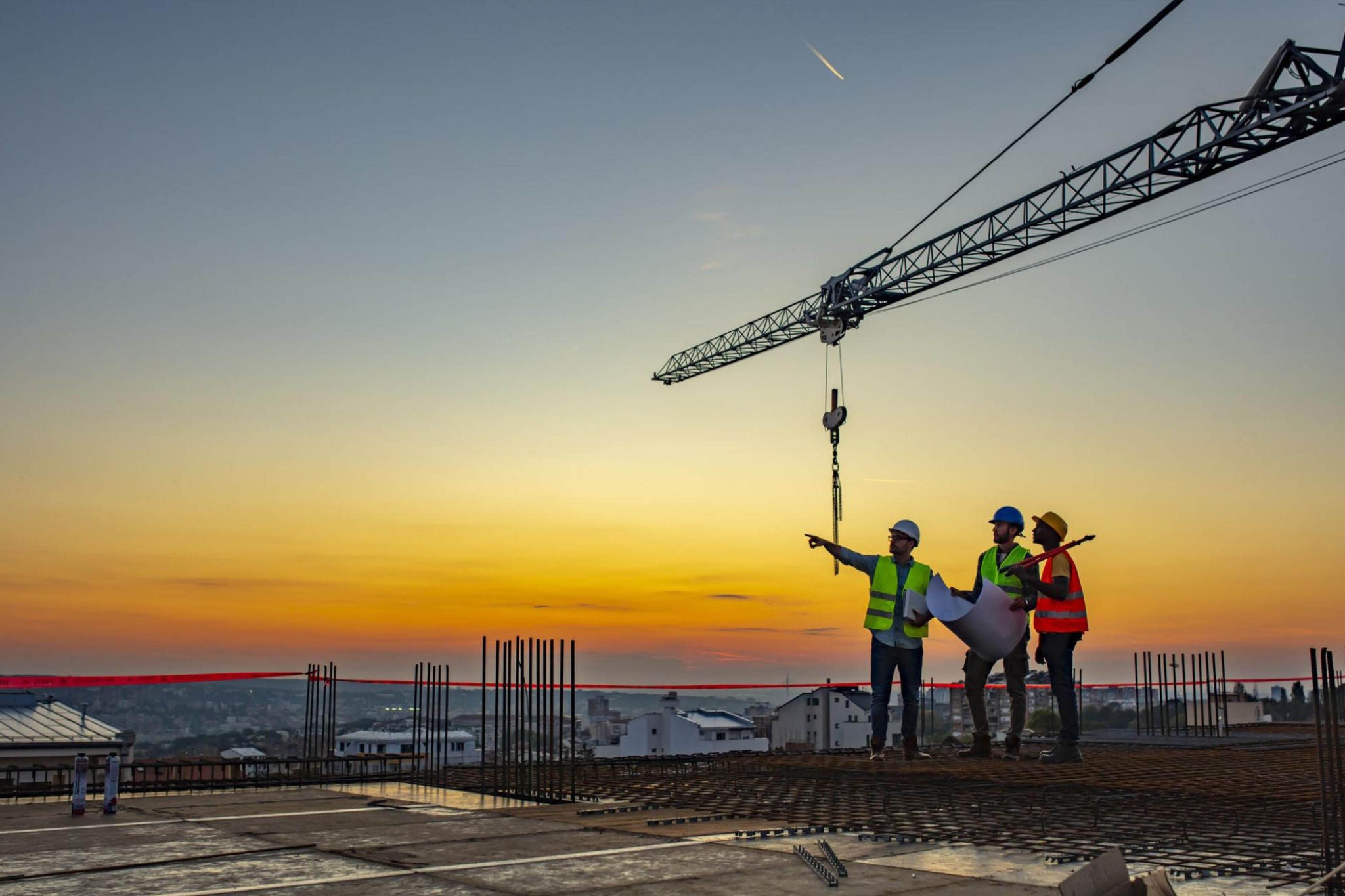 People examining project plans on a construction site with a tower crane and urban skyline in the background.