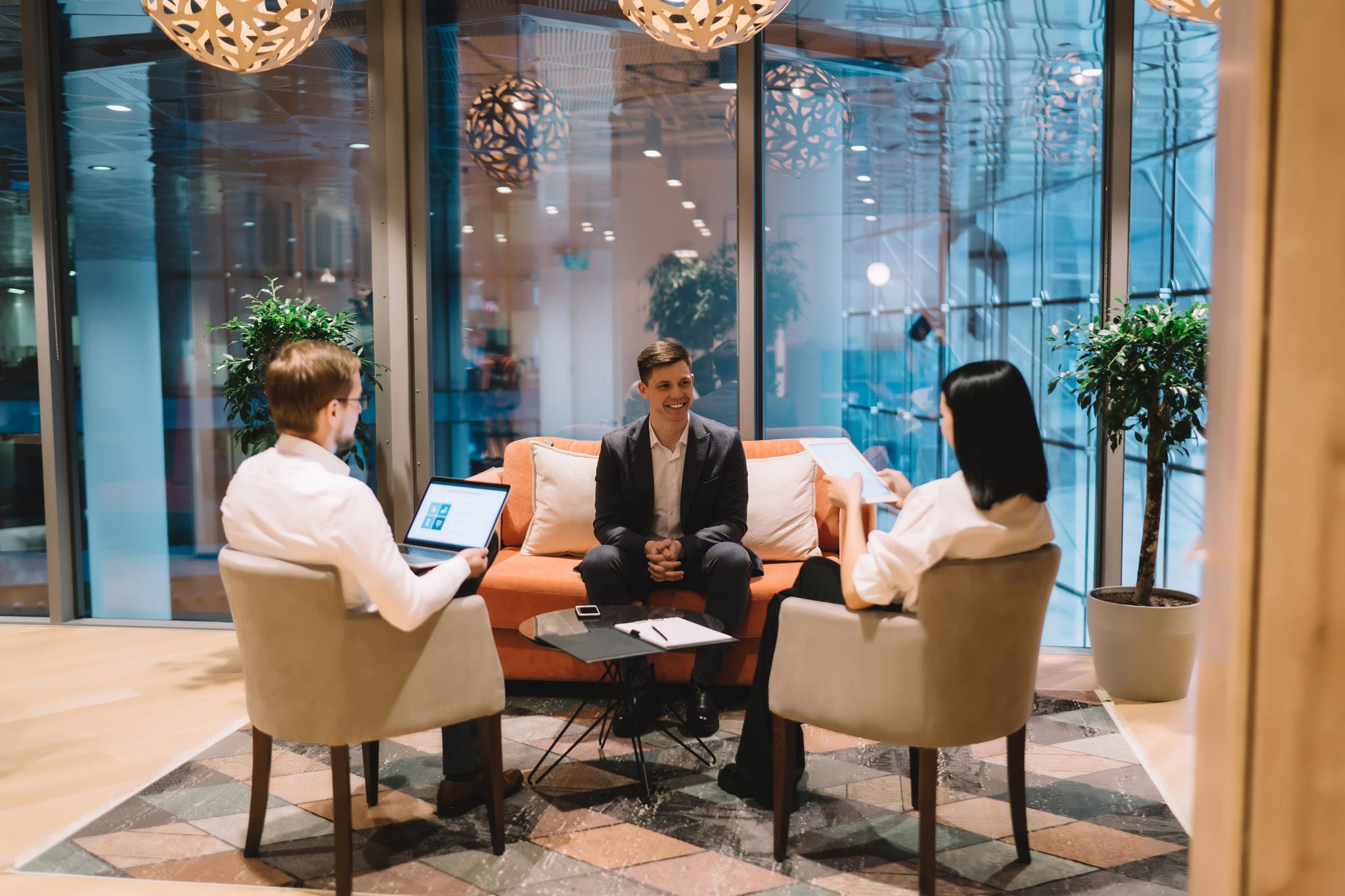 Managers of HR department using laptop and tablet while interviewing confident job applicant in formal black suit during hiring process in sitting area of modern office