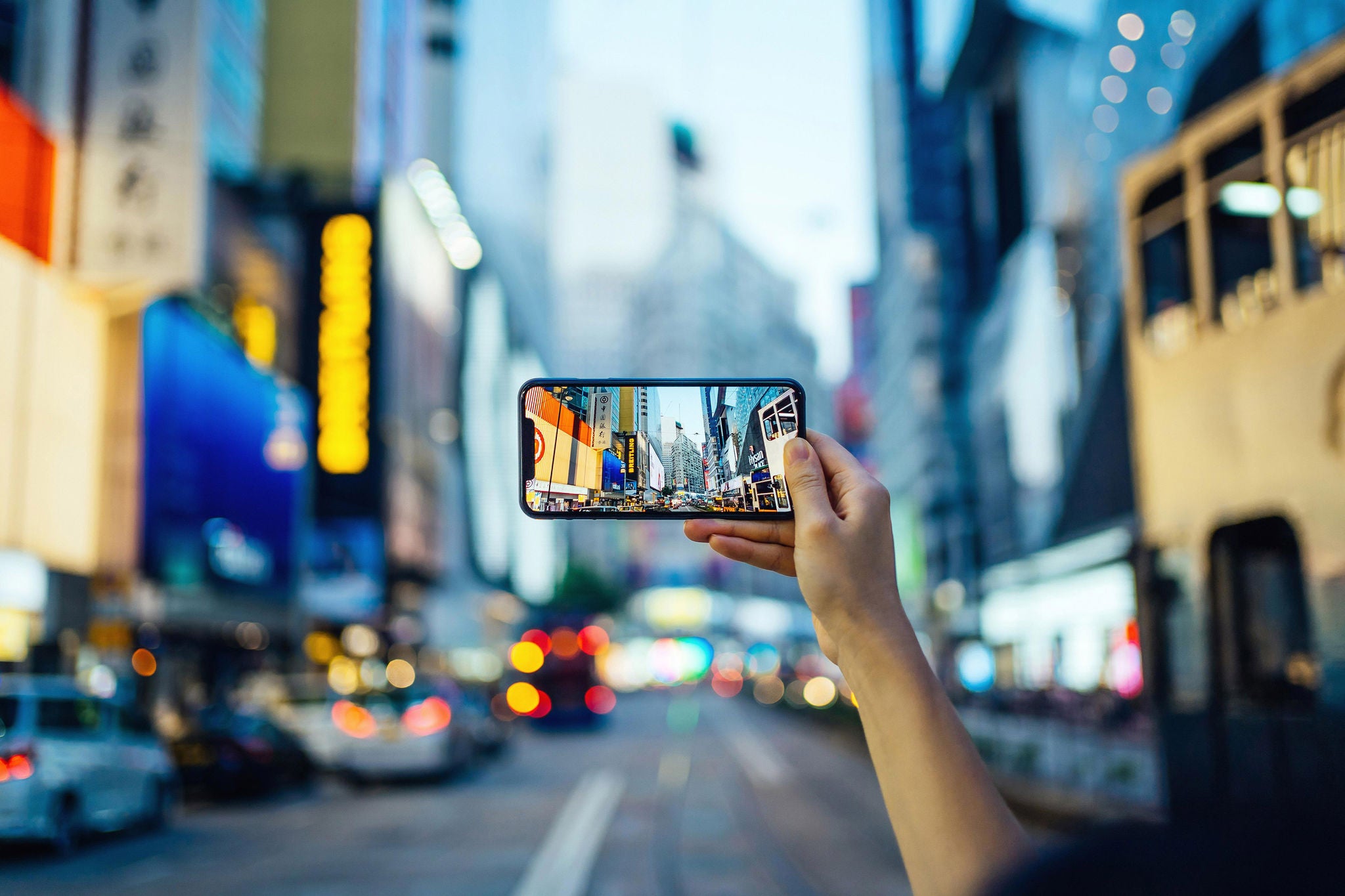 Woman's hand taking photo with smartphone in busy downtown district against urban skyscrapers with multi-coloured neon signs and city traffic