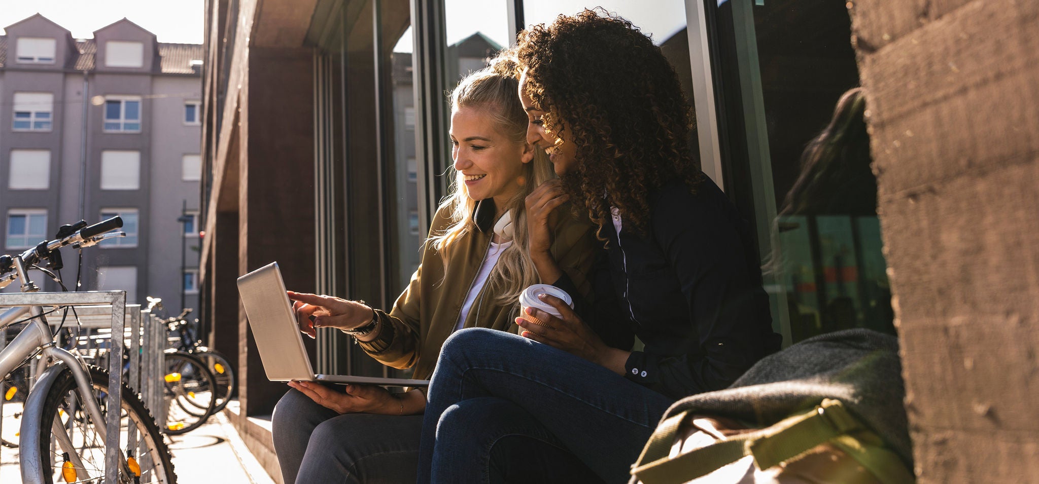 Zwei Personen sitzen draußen auf einer Bank, betrachten einen Laptop und trinken Kaffee, sonnige Szene