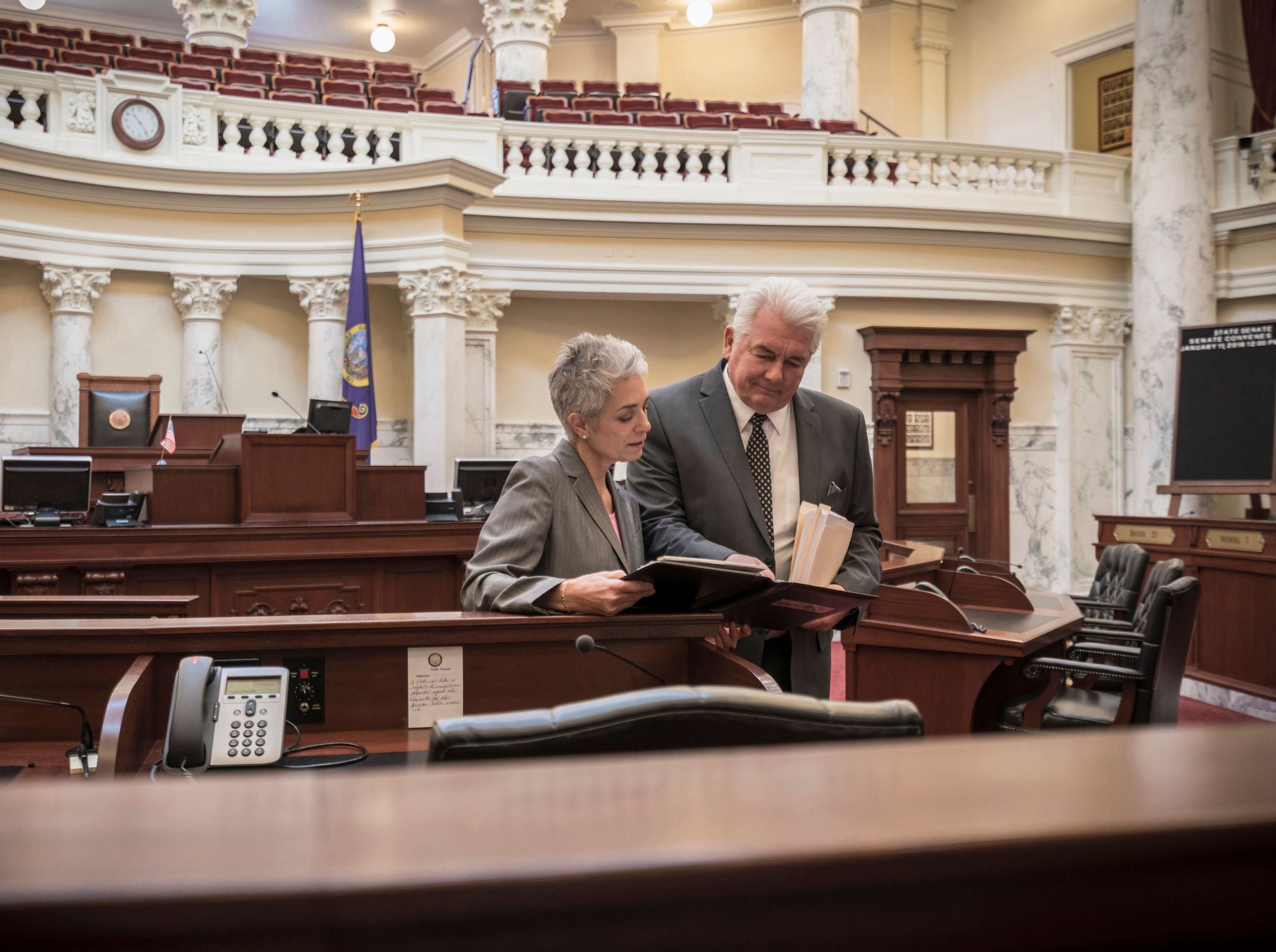 Egy díszes, történelmi jellegű ülésteremben két felnőtt személy áll egy padsor mellett, miközben dokumentumokat és iratokat néznek át.