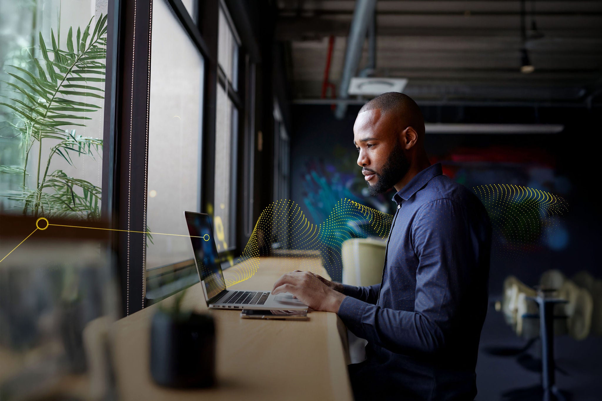 Mid adult black male creative sitting by window in an office social area using a laptop, side view