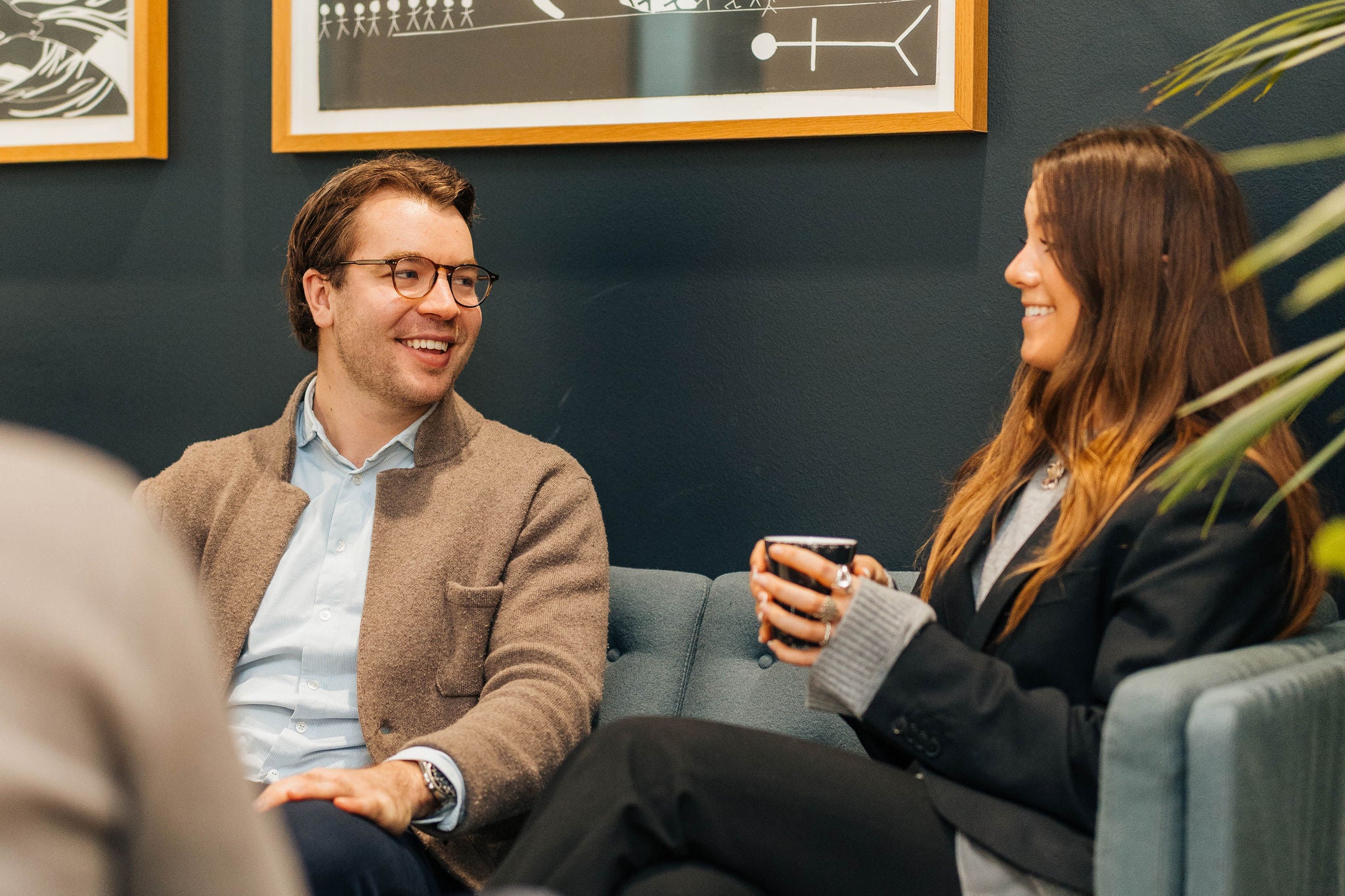 Two colleagues sitting on a sofa, talking and holding cups in a relaxed office lounge.