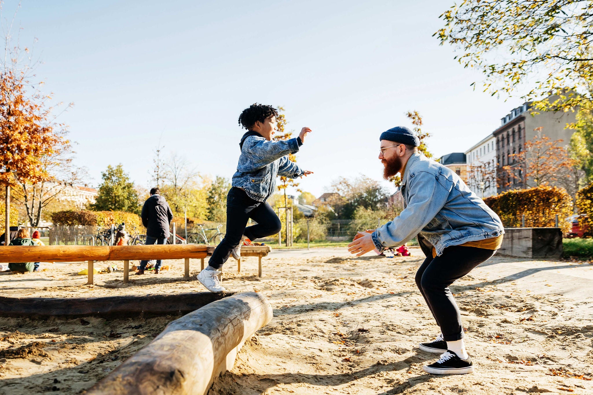 A young boy leaping into his fathers arms from a log while messing around in a playground at the park together.