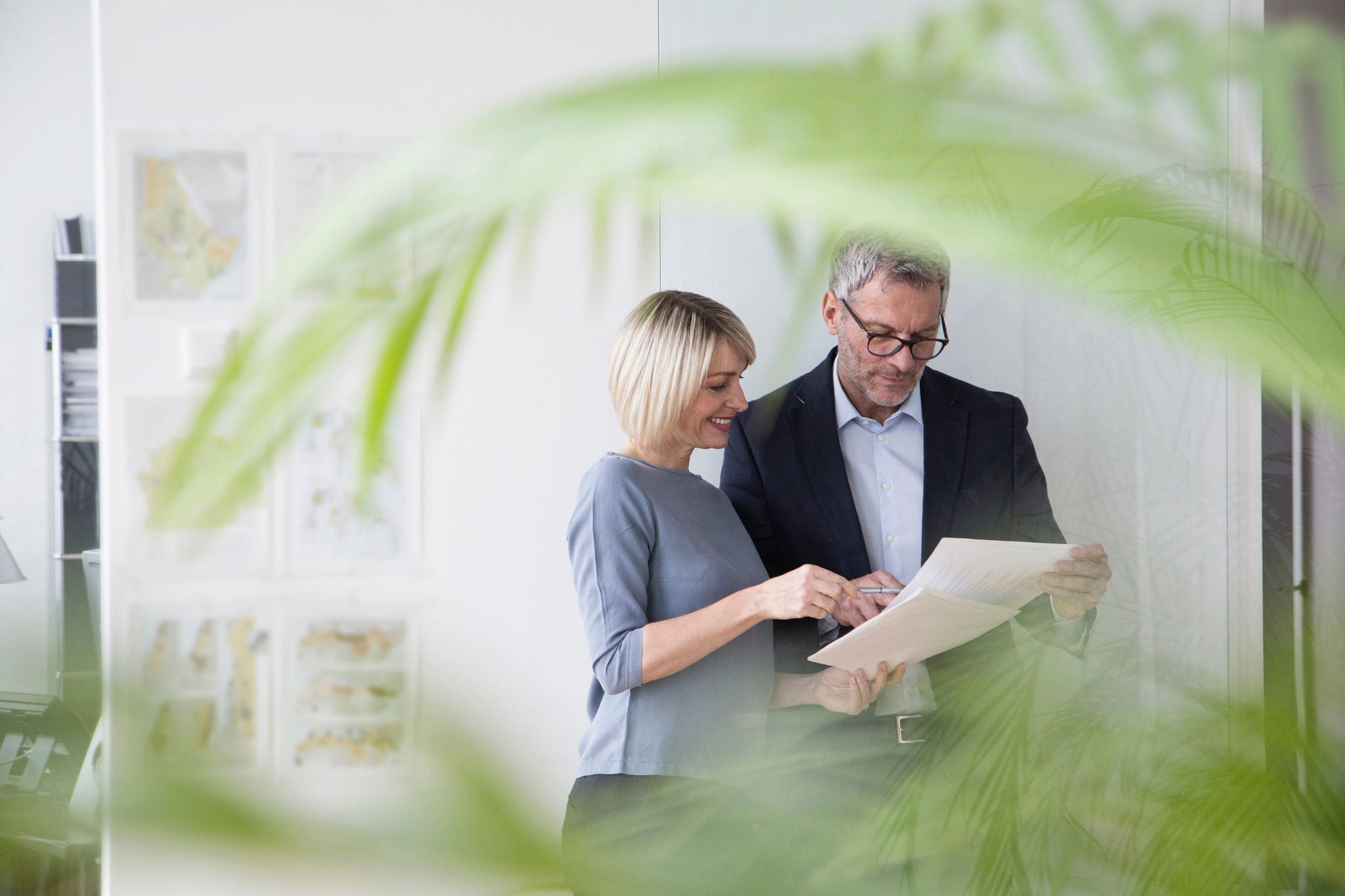 Två kollegor på kontor granskar dokument tillsammans, sedda genom gröna växter