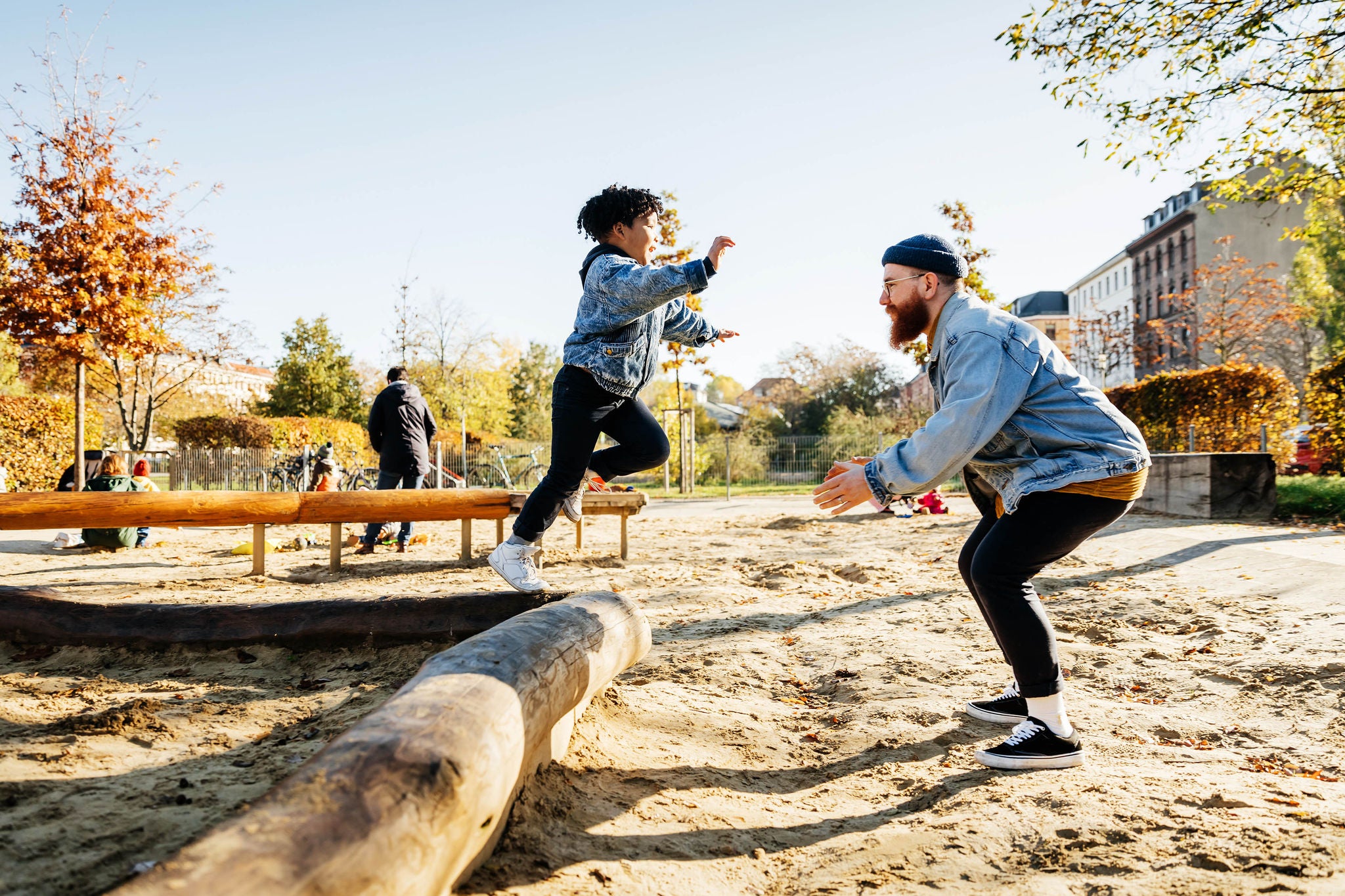 A young boy leaping into his fathers arms from a log while messing around in a playground at the park together.