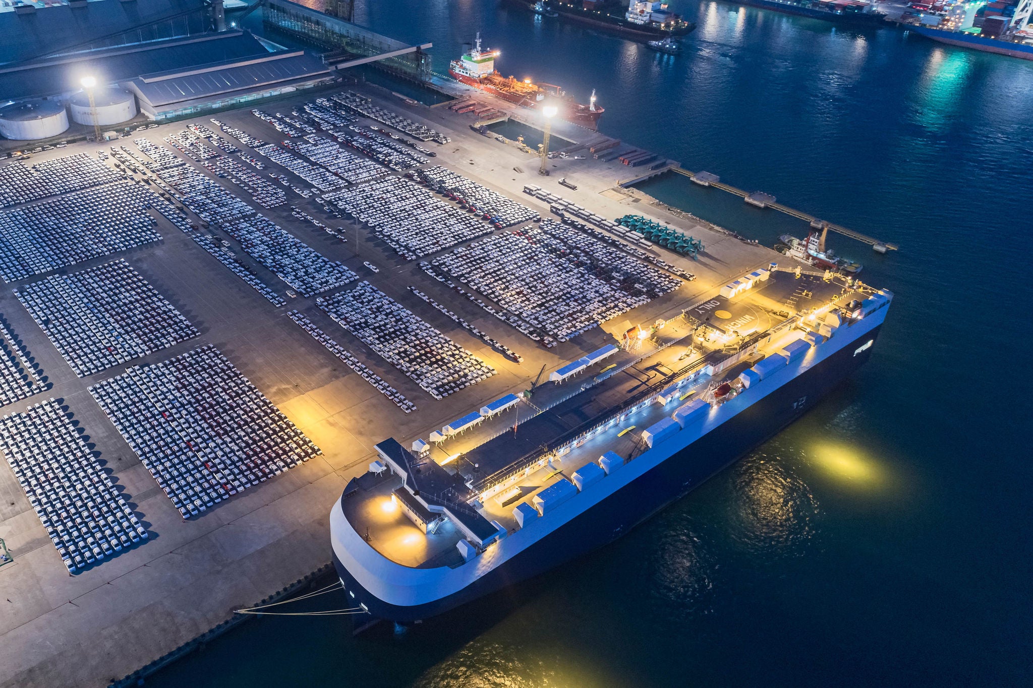 Luchtbeeld van auto’s opgesteld in een haven naast een groot vrachtschip ’s nachts