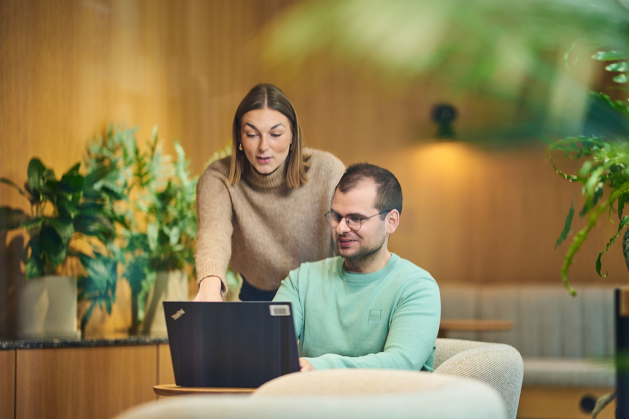 Two colleagues reviewing information on a laptop in a modern office lounge with plants and wooden interiors.