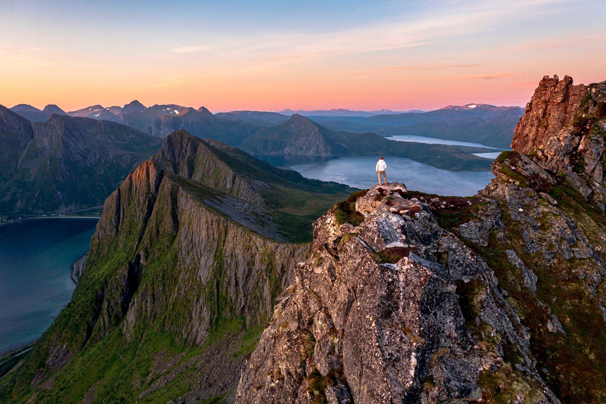 En person står på en fjelltopp med utsikt over en fjord og dramatiske fjellformasjoner i norsk landskap i solnedgang.