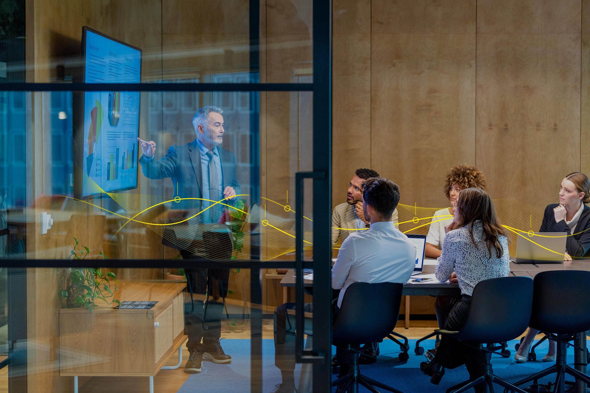 Mature man giving a big data presentation on a tv in a board room. There are several financial graphs and charts on the screen with a diverse group of people in the meeting room. There is paperwork and technology on the table