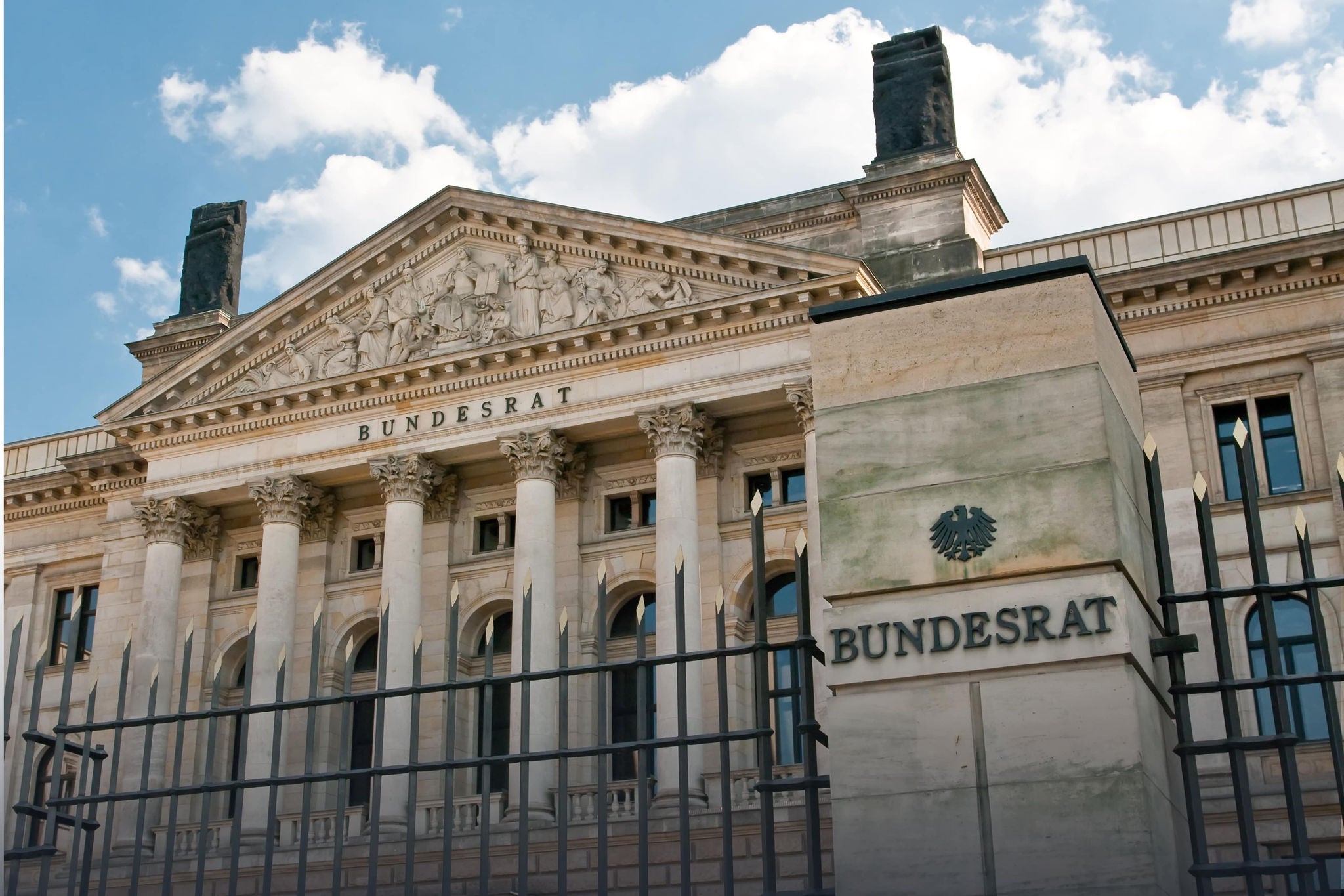 A smiling young woman with headphones and folders, standing outdoors on a college campus.