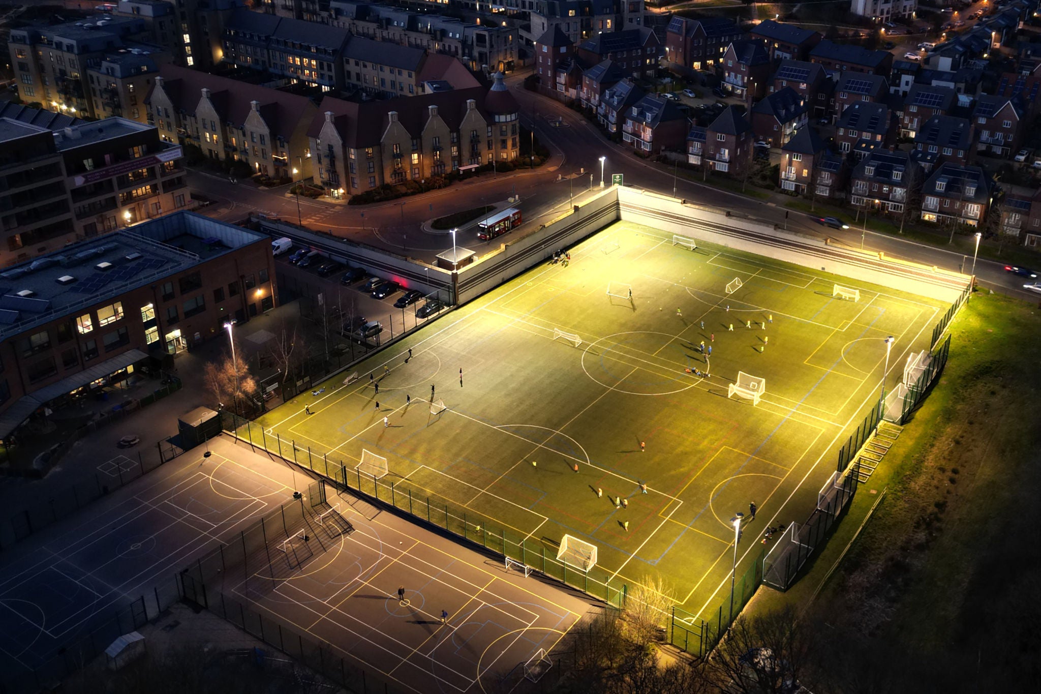 Aerial view of a floodlit sports pitch and outdoor courts surrounded by residential buildings in an urban neighbourhood at dusk.