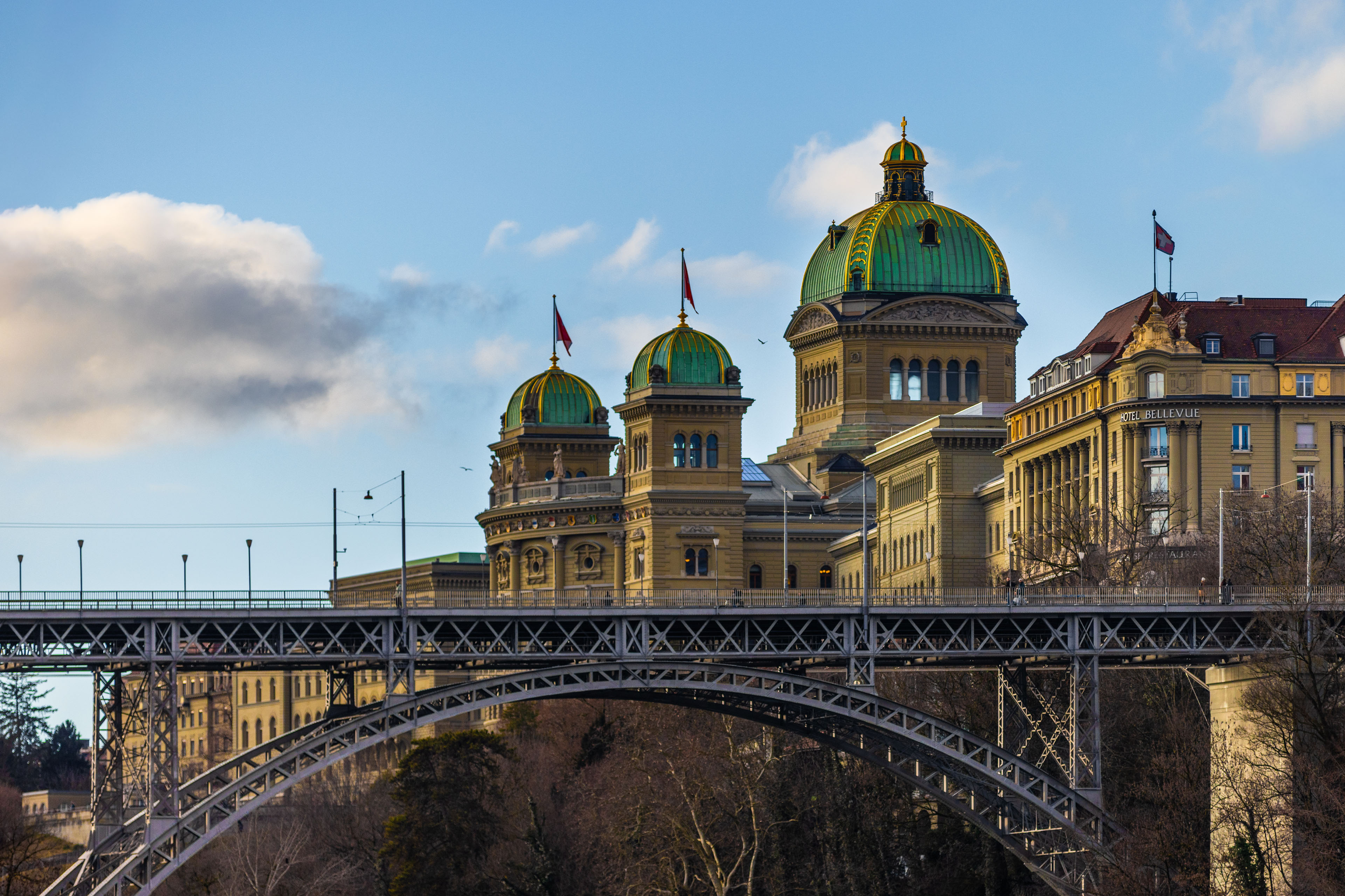 Historic cityscape featuring the Swiss Federal Parliament Building with its distinctive green copper domes and Swiss flags, viewed behind an arched steel bridge under a blue sky with scattered clouds.