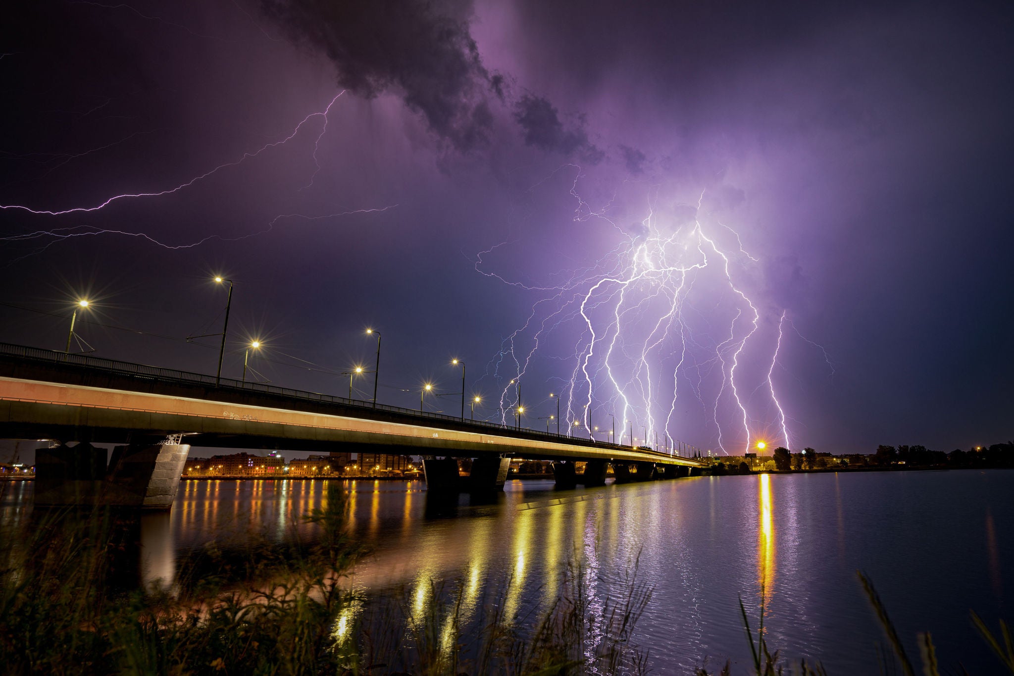 Mehrere Blitze erhellen den Himmel über einer beleuchteten Brücke und spiegeln sich im Wasser.