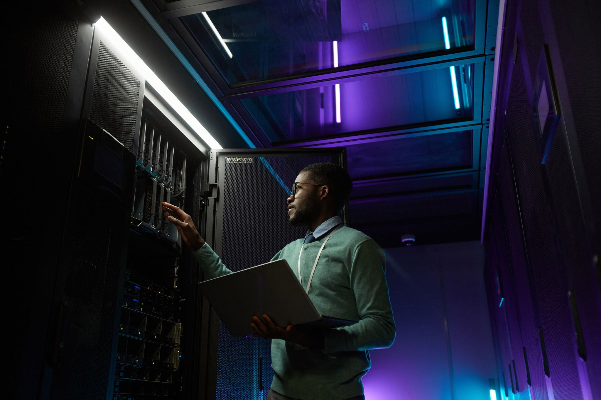 Low angle portrait of young African American data engineer working with supercomputer in server room lit by blue light