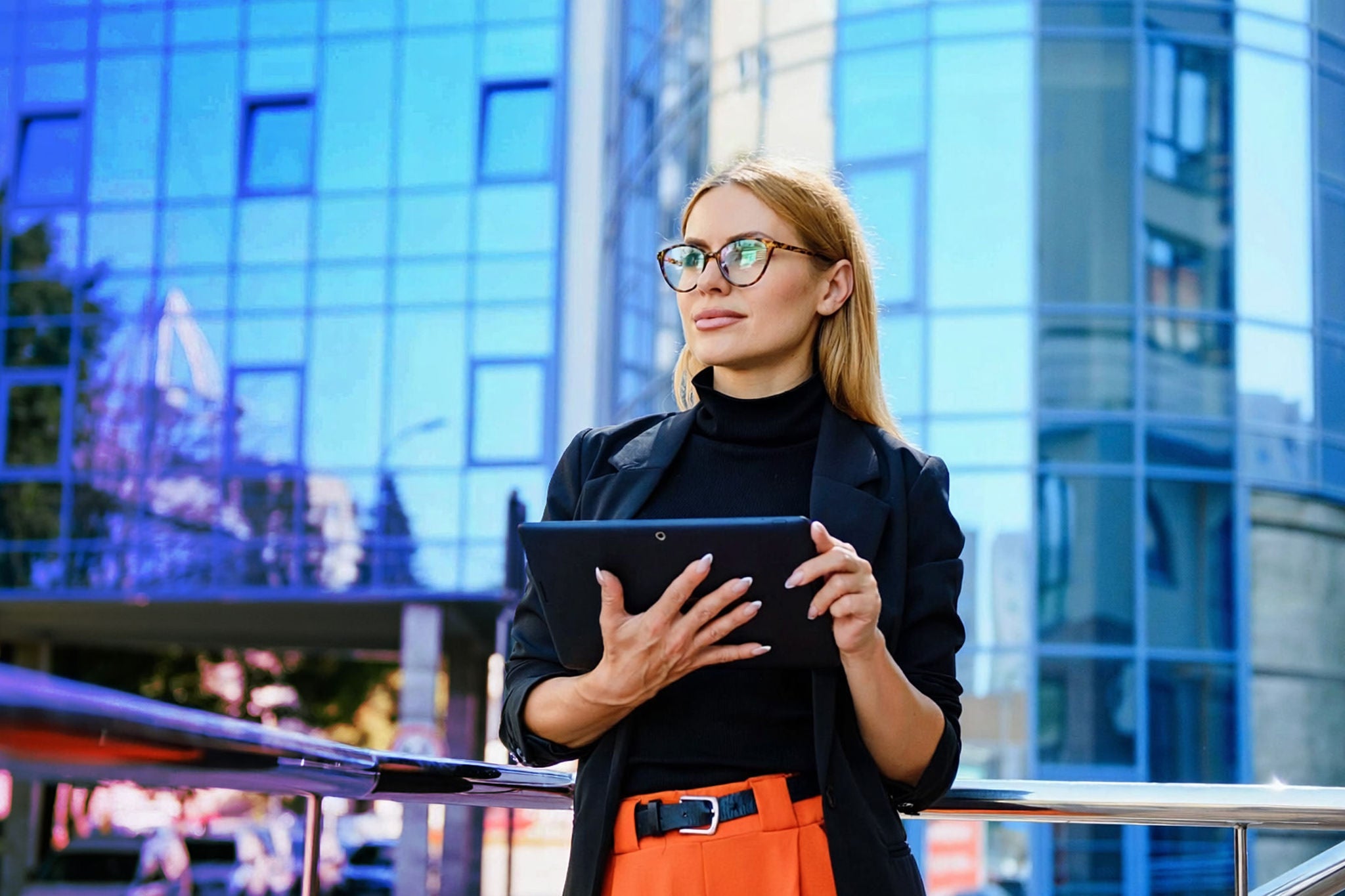 Female banker using a tablet and wireless earphones outdoors near her office at dusk.