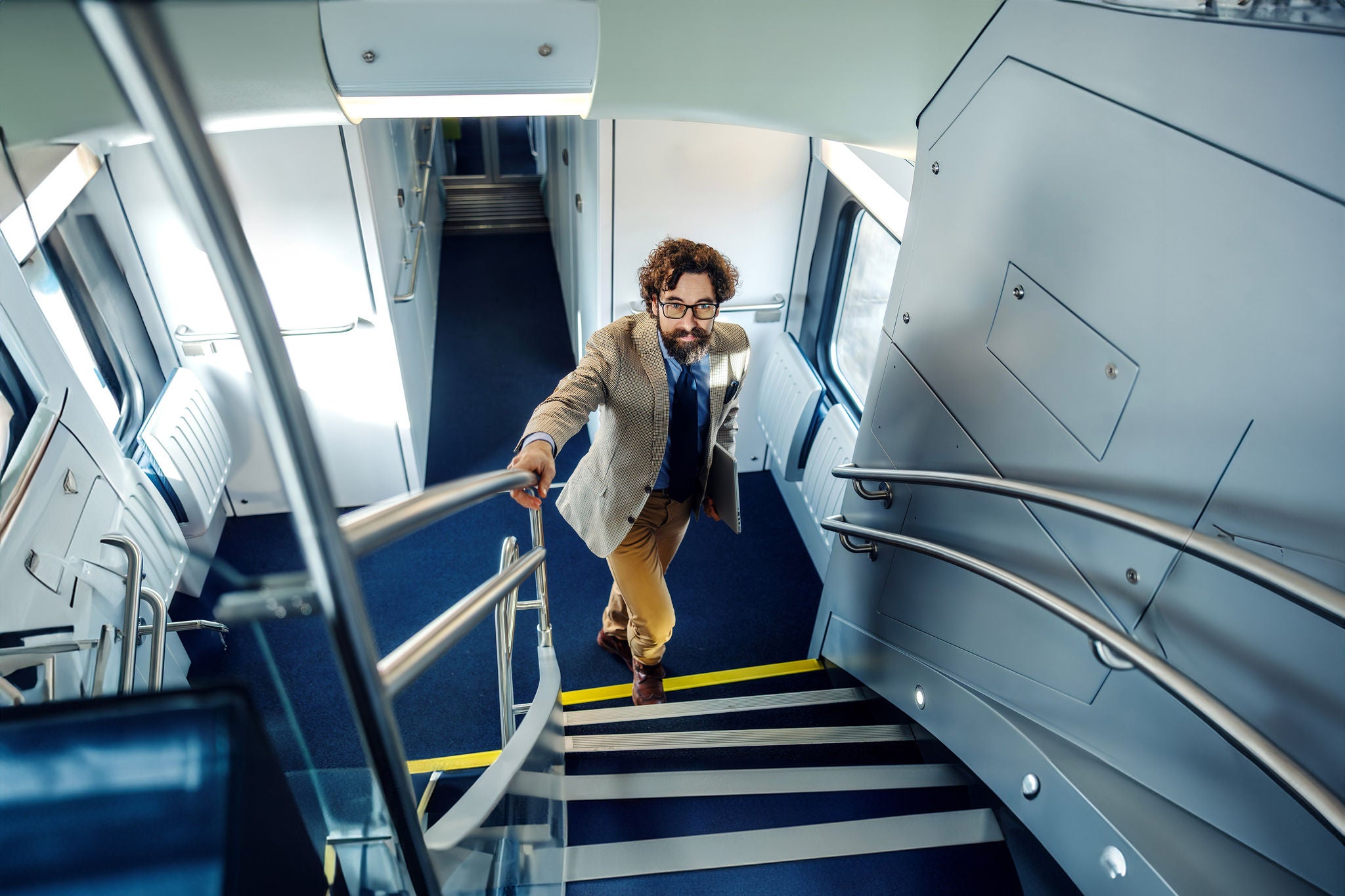 Man in business casual attire walking up stairs inside a modern train, holding a laptop in one hand and a railing with the other, traveling for business