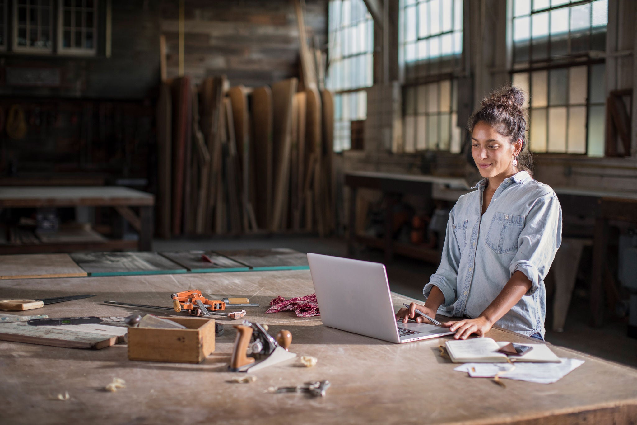 Young Mixed Race Female Entrepreneur Solving a Complicated Business Challenge with Pencil, Laptop, Carpentry Tools, and Confidence. Young mixed race woman stands at her sturdy wooden worktable, strewn with carpentry tools, sawdust, paper, and other creative mayhem. She's using a laptop and also has a smartphone on the tabletop. Innovation is evident in her confident calm posture and the sparks of discovery/innovation lighting up her half-smiling face. In the background, natural light of the spacious, lofty maker space illuminates raw lumber and other woodworking tools in soft focus.