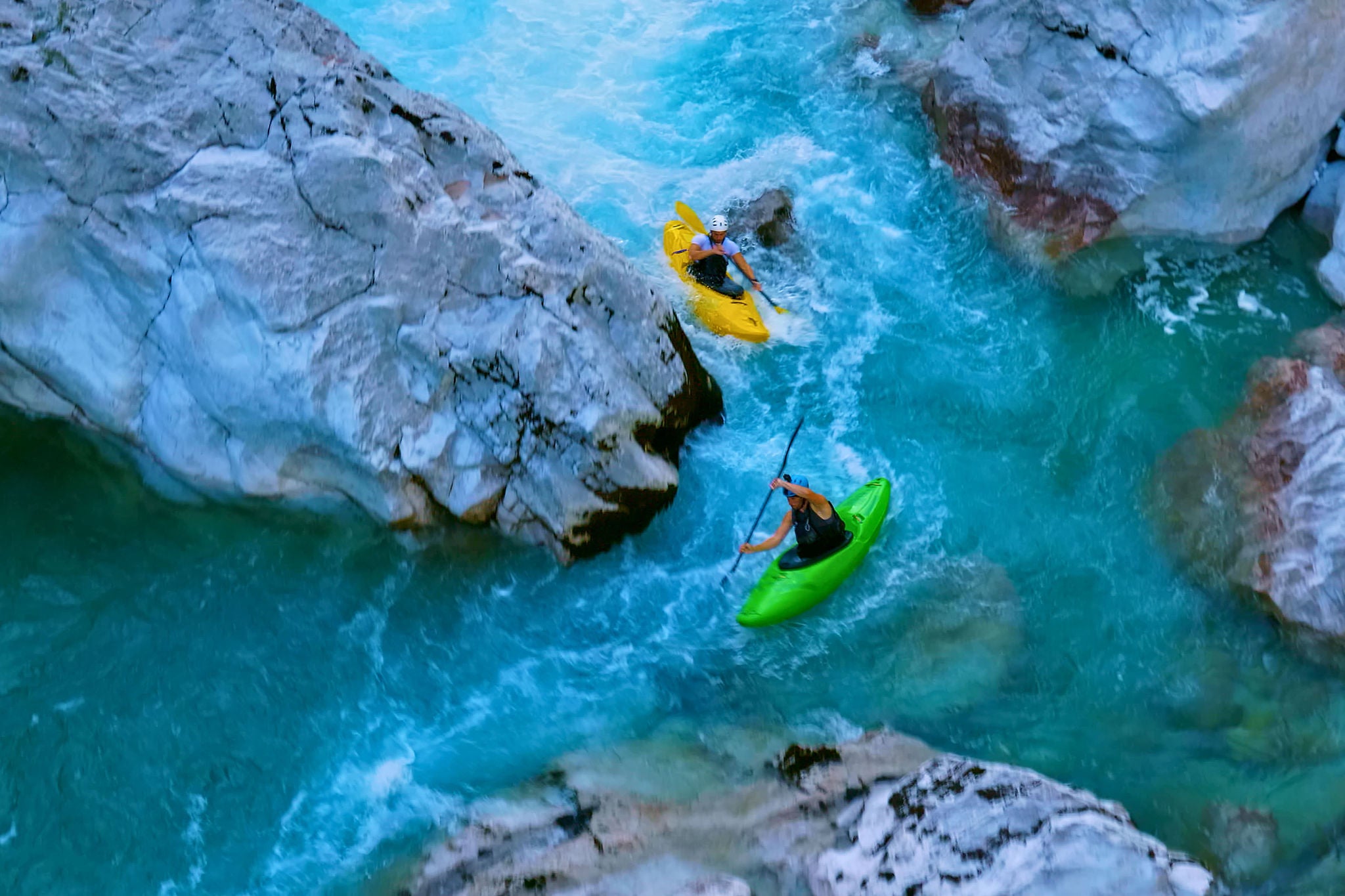 One kayaker, wearing a white helmet, paddles a yellow kayak, while the other, in a black.