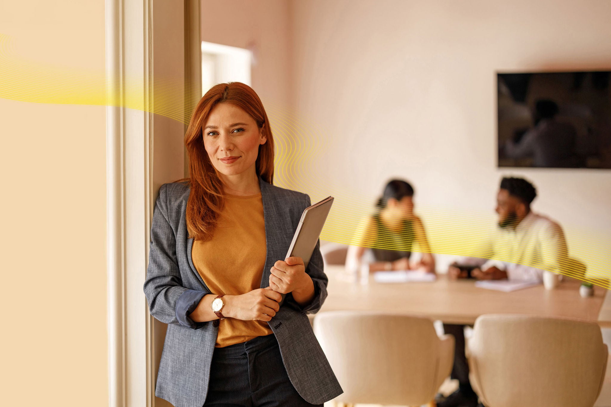 Confident female manager holding laptop leaning on wall with team of colleagues collaborating in background