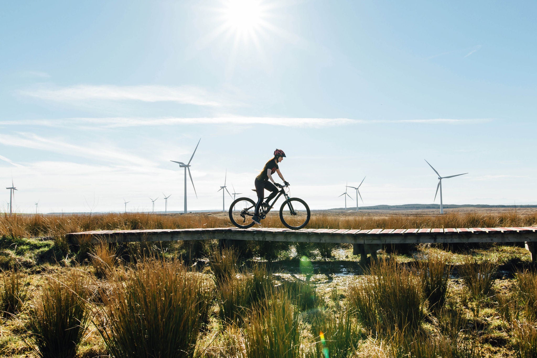A woman riding a mountain bike in over a wooden track surrounded by wild grass at a wind farm in Scotland