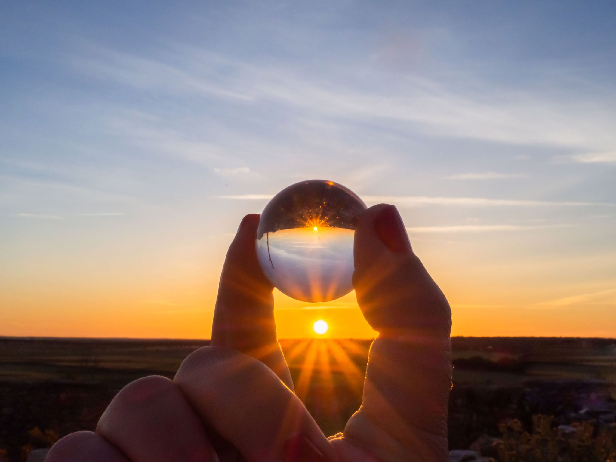 Egy kézen tartott üveggolyón keresztül látszik a lenyugvó nap, amely a horizont felett ragyogva a gömbben tükröződik.
