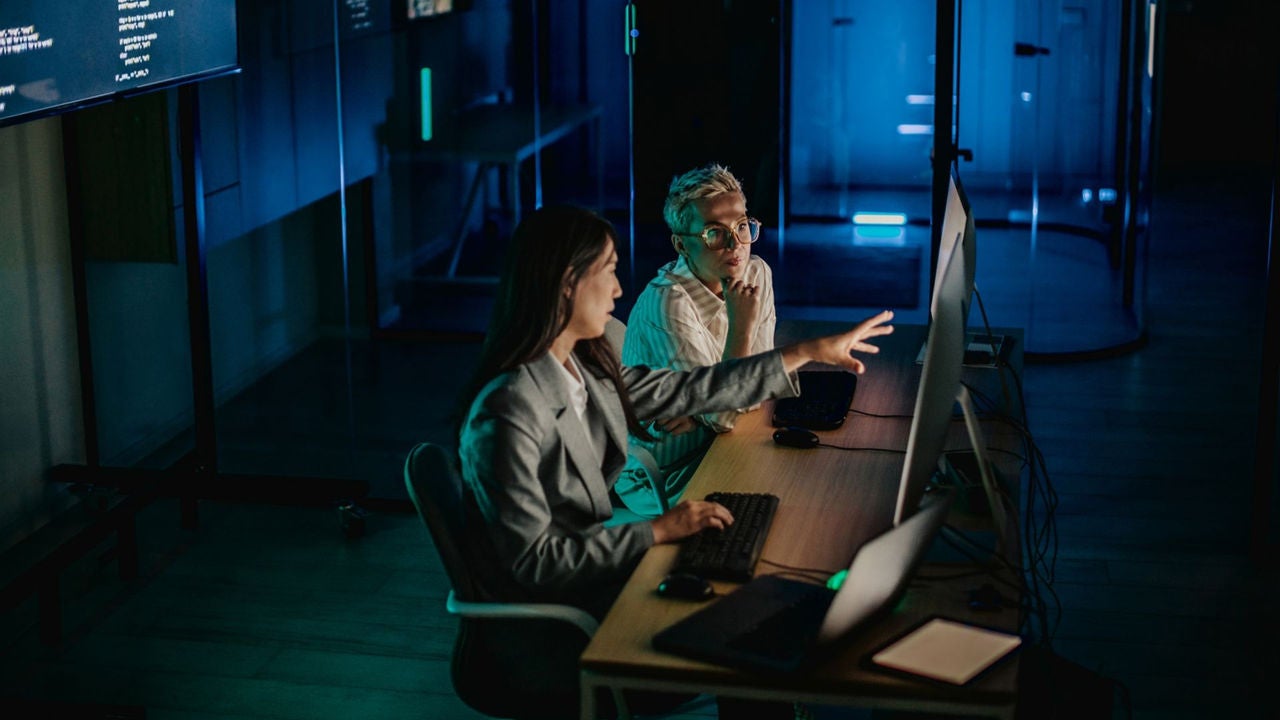 Two adults work together in a bright data center, focusing on screens as they analyze data and develop software solutions.