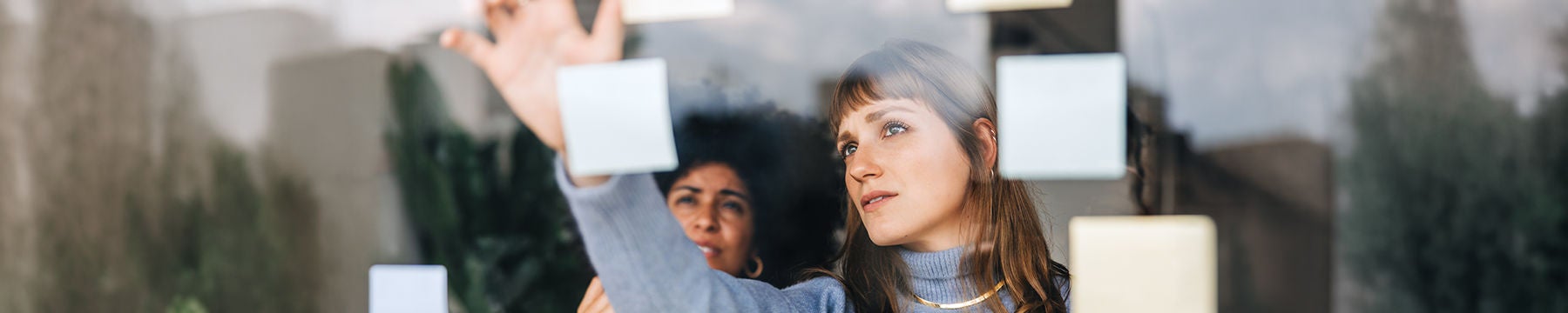 Mitarbeitende organisieren Ideen mit Haftnotizen an einer Glaswand im Büro. Kollaborative Planung im Business‑Kontext.