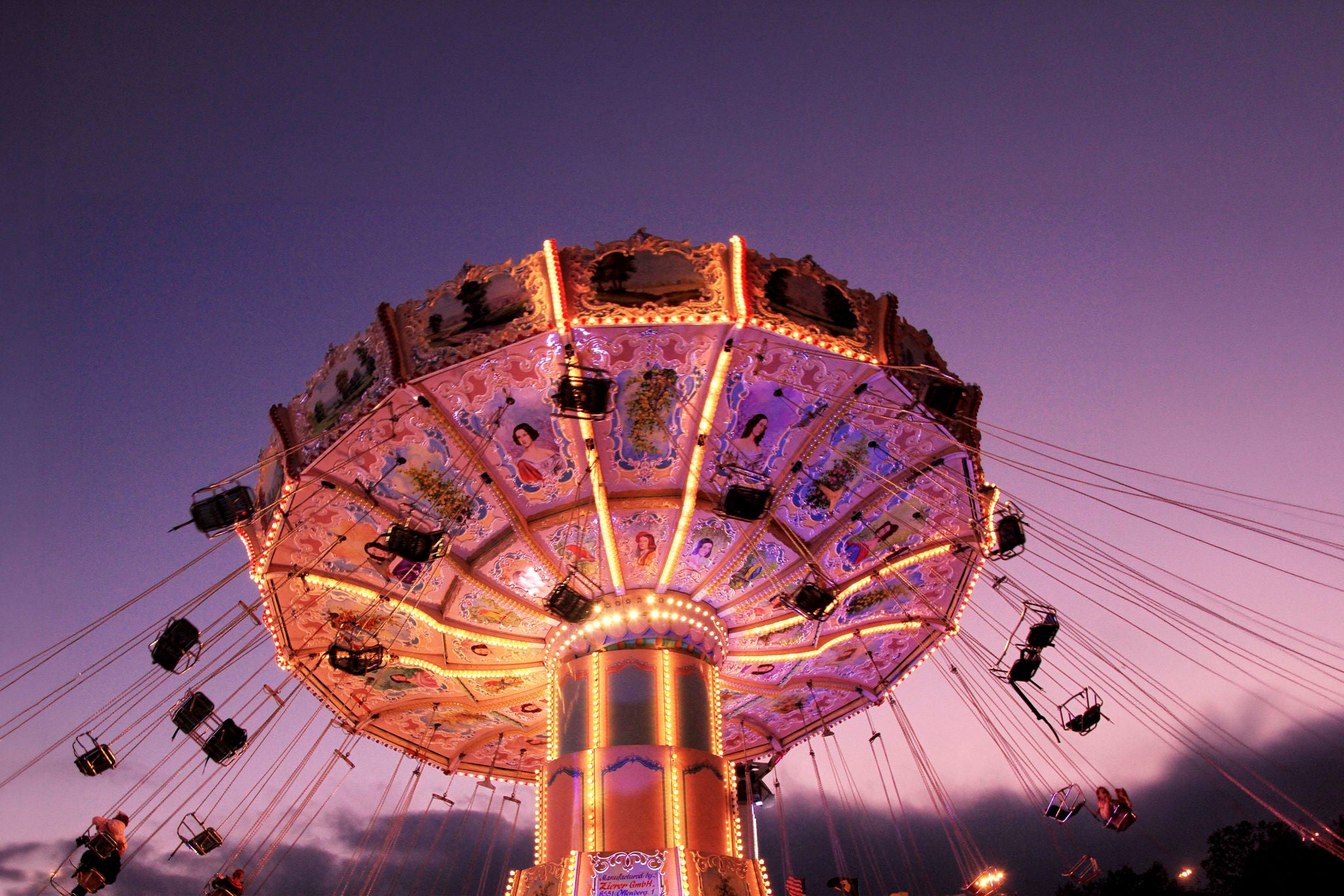 Illuminated swing carousel ride at dusk, with suspended chairs and decorative lights against a twilight sky