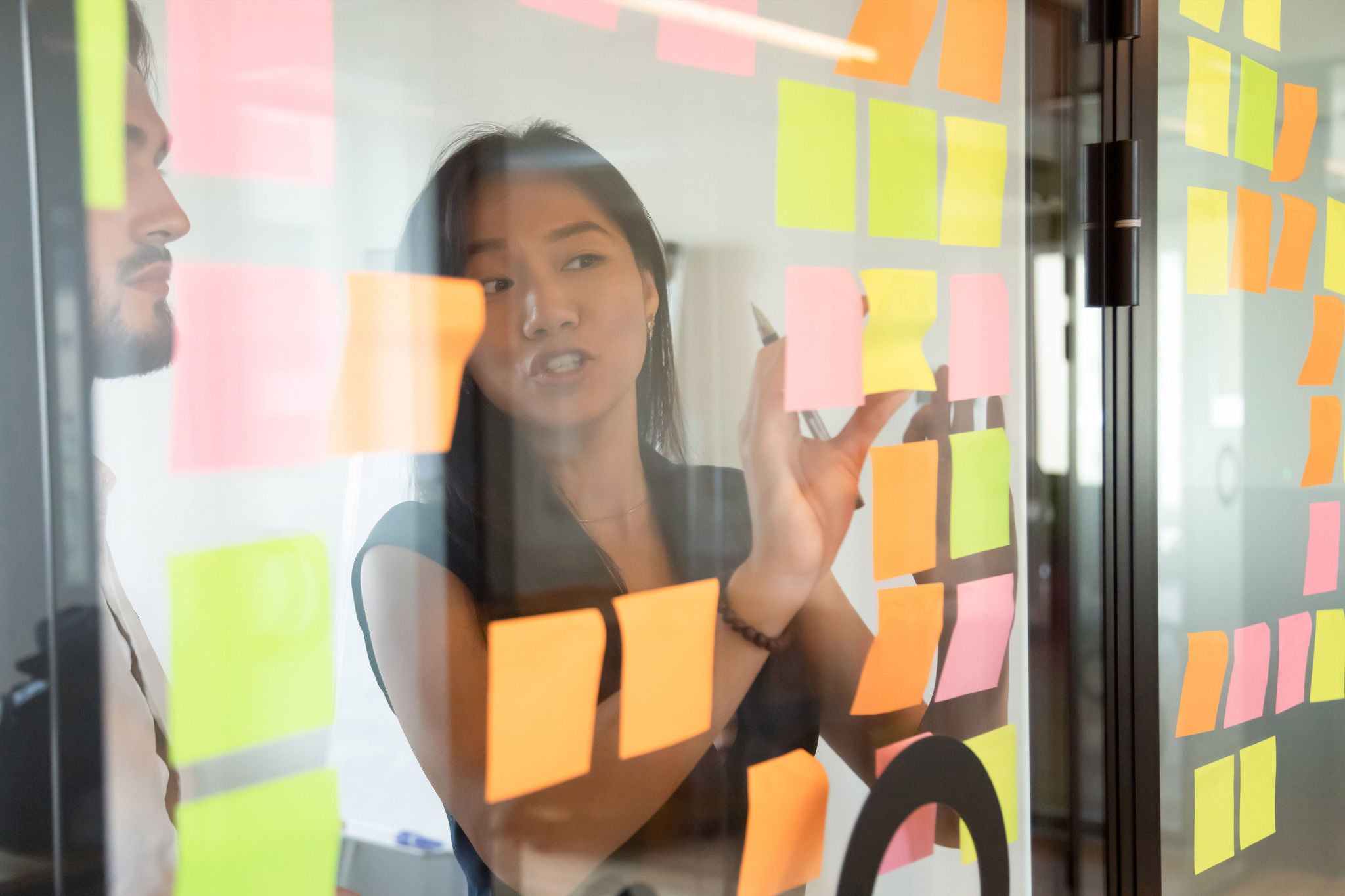 View through glass partition asian female worker working strategizing with caucasian male colleague, attached colourful post-it notes on glass wall where written ideas actions plan for aim achievement