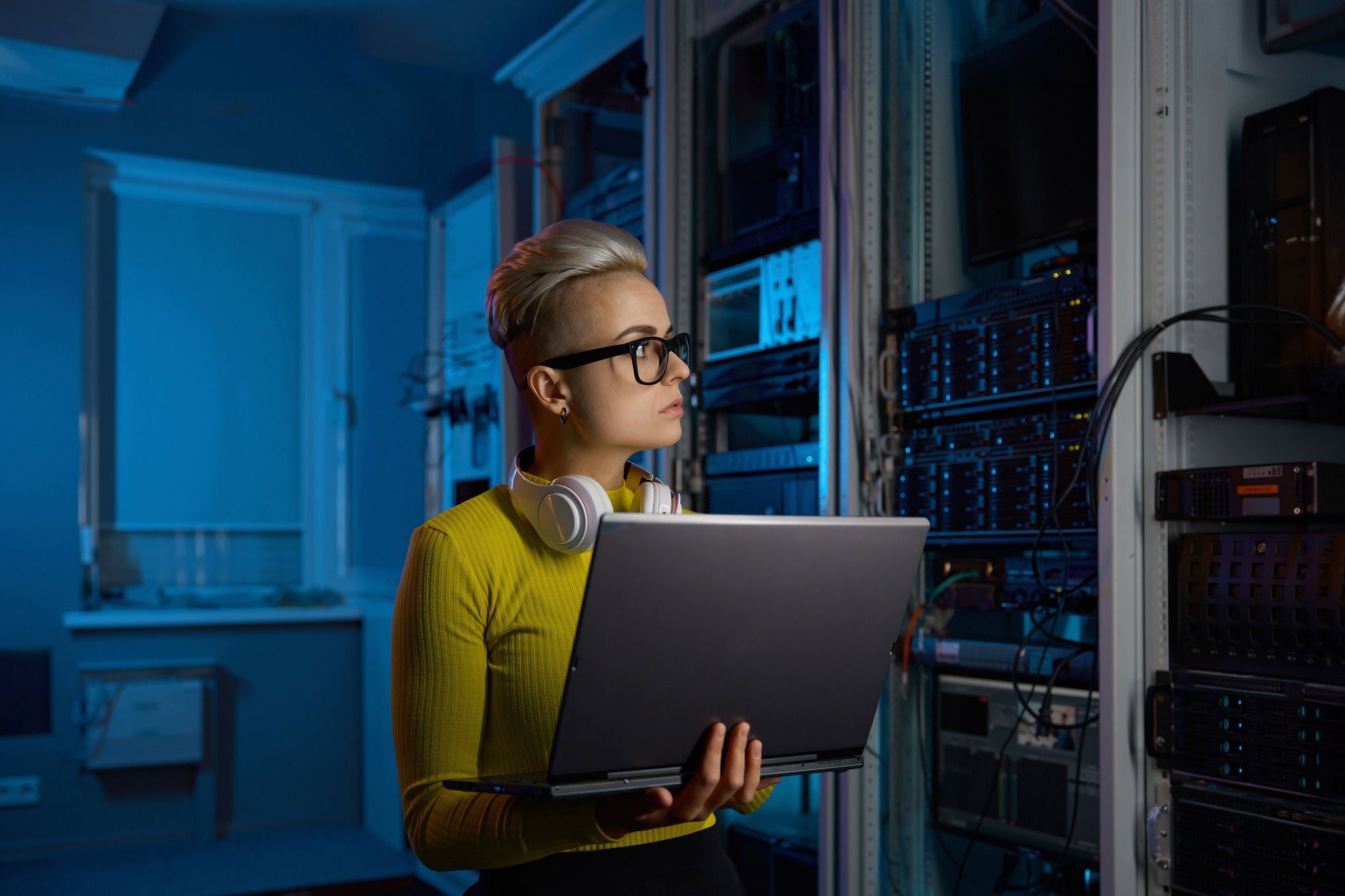 Portrait of concentrated young woman coder solving problem or troubleshooting using laptop in dark server room of modern data center
